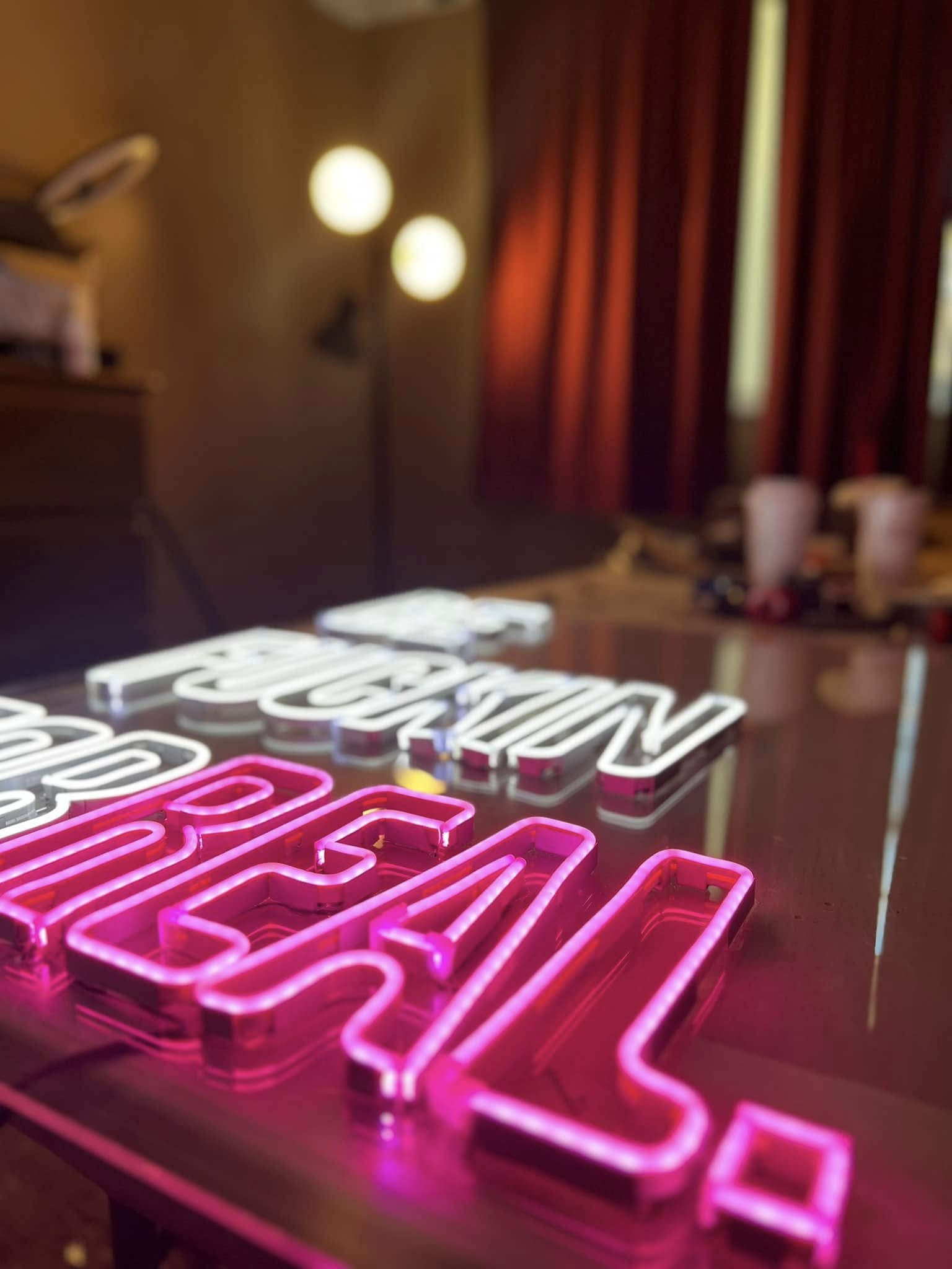 Close-up of illuminated neon pink and white sign with partially visible text in the foreground. Red curtains, a floor lamp, and blurred lights are visible in the background.