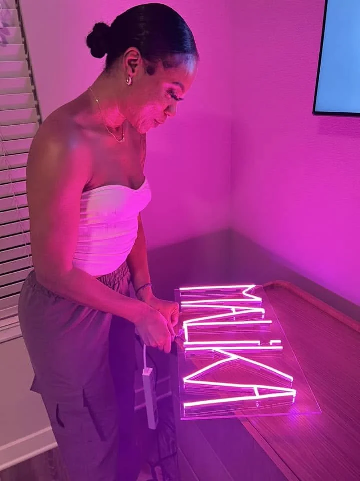 A woman is holding a pink neon sign that spells 'MALIK' in a room with pink lighting, standing next to a wooden table.