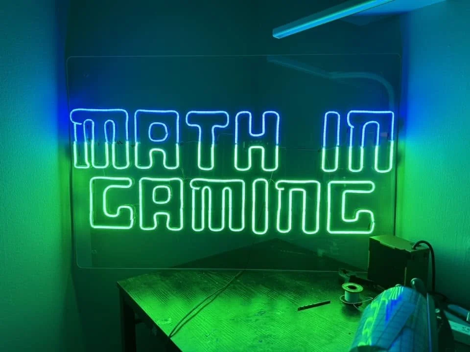 Neon sign reading 'MATH 100 GAMING' in blue and green colors, with a wooden desk below and some electronic items on it.