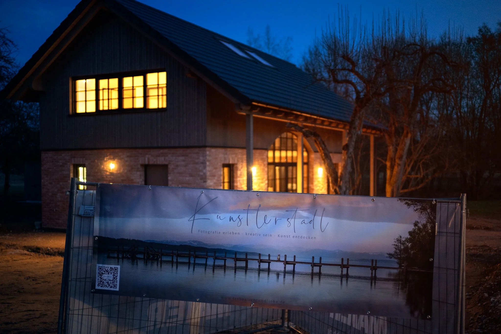 Ein modernes Haus bei Dämmerung mit beleuchteten Fenstern, im Vordergrund ein Bauzaun mit einem Banner für die Kunststädte-Ausstellung.
