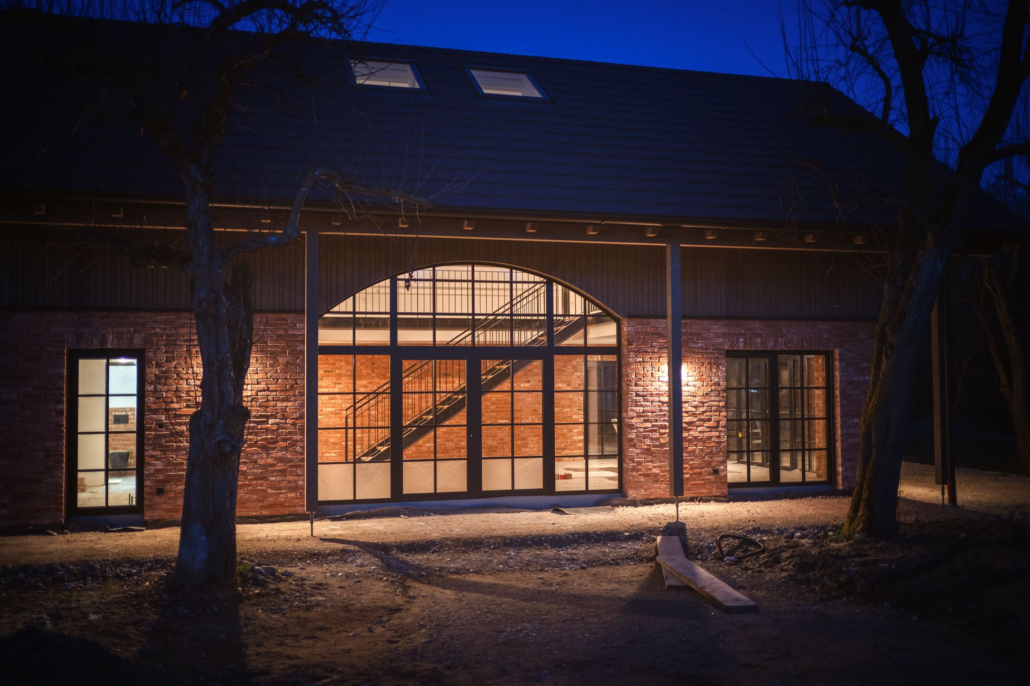 Ein modernes Haus mit großen Glasfenstern und Ziegelwand, beleuchtet bei Nacht, umgeben von Bäumen.