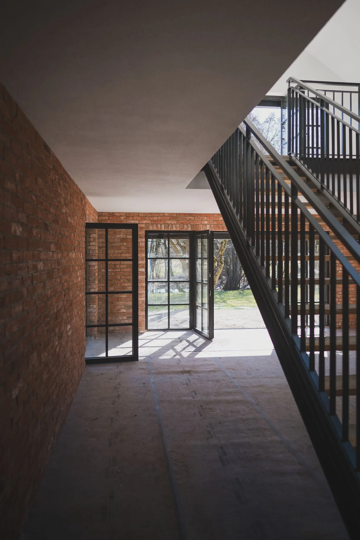Innenraum eines modernen, unfertigen Gebäudes mit roten Ziegelwänden, schwarzen Metallfenstern und -türen, einer Treppe mit schwarzem Geländer und Blick nach draußen auf eine grüne Landschaft.