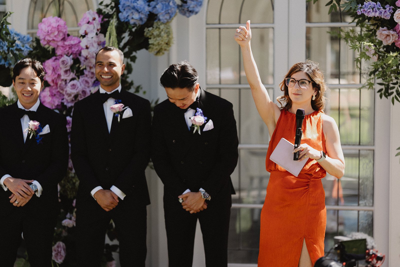 a woman in an orange dress stands with 3 men dressed in suits in front of a floral arbour.