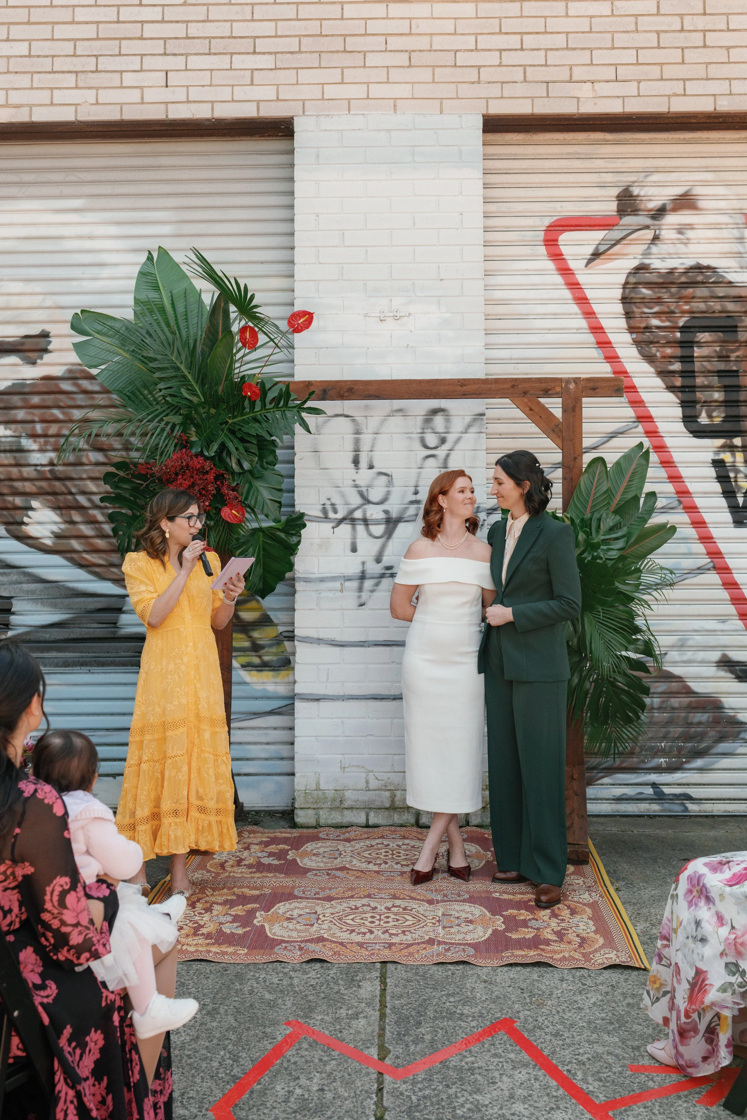 celebrant in yellow dress stands in front of colourful background with two brides one wearing a white dress and the other in a dark green suit