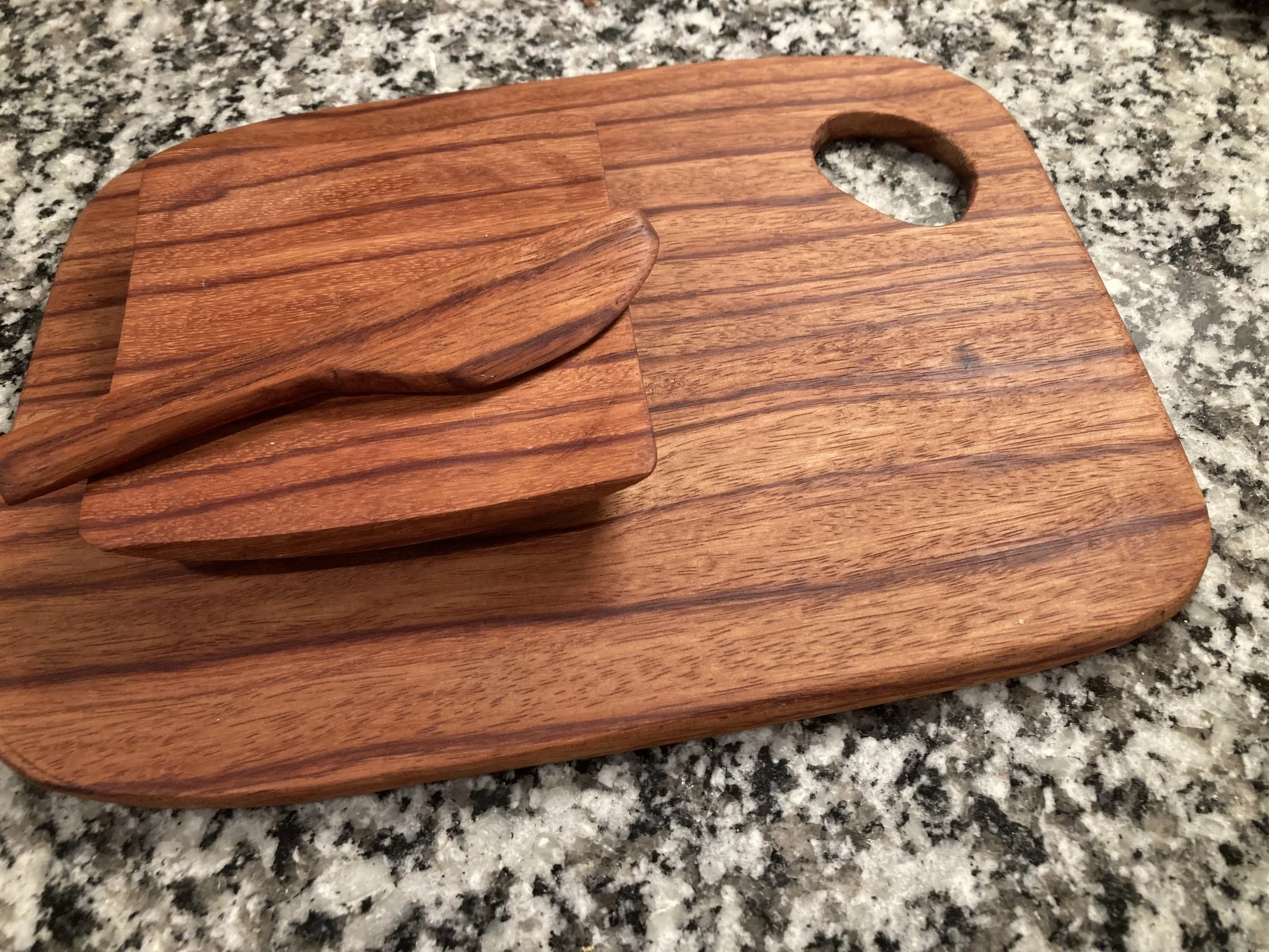 A wooden cutting board with a handle, featuring two matching wooden knives, resting on a speckled granite countertop.