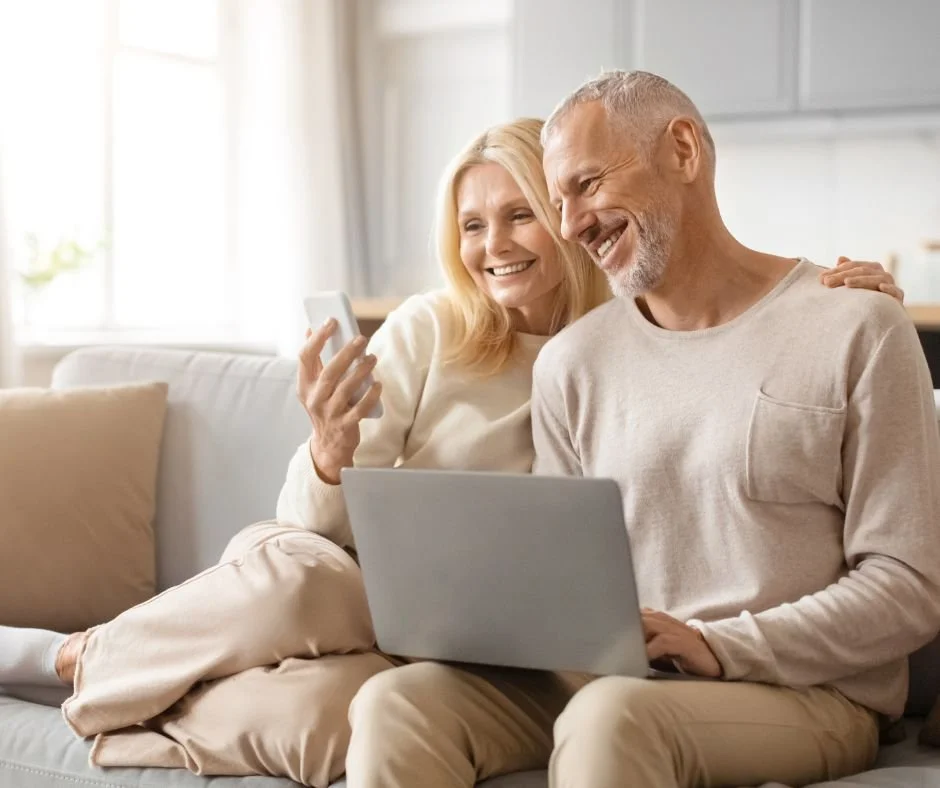 An older couple sitting on a beige sofa in a bright living room, looking at a laptop and a smartphone, smiling and enjoying their time together.