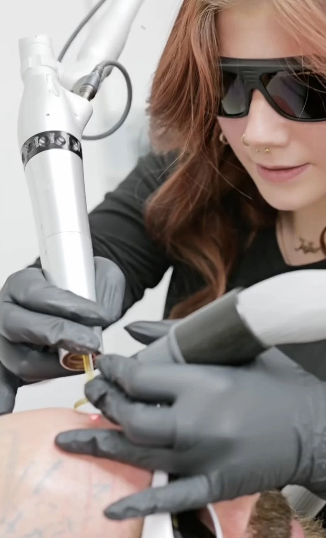 A woman with tattooed arms and wavy hair is sitting at a table, tattooing on someone's arm. She is wearing black gloves and focused on her work in a tattoo studio.