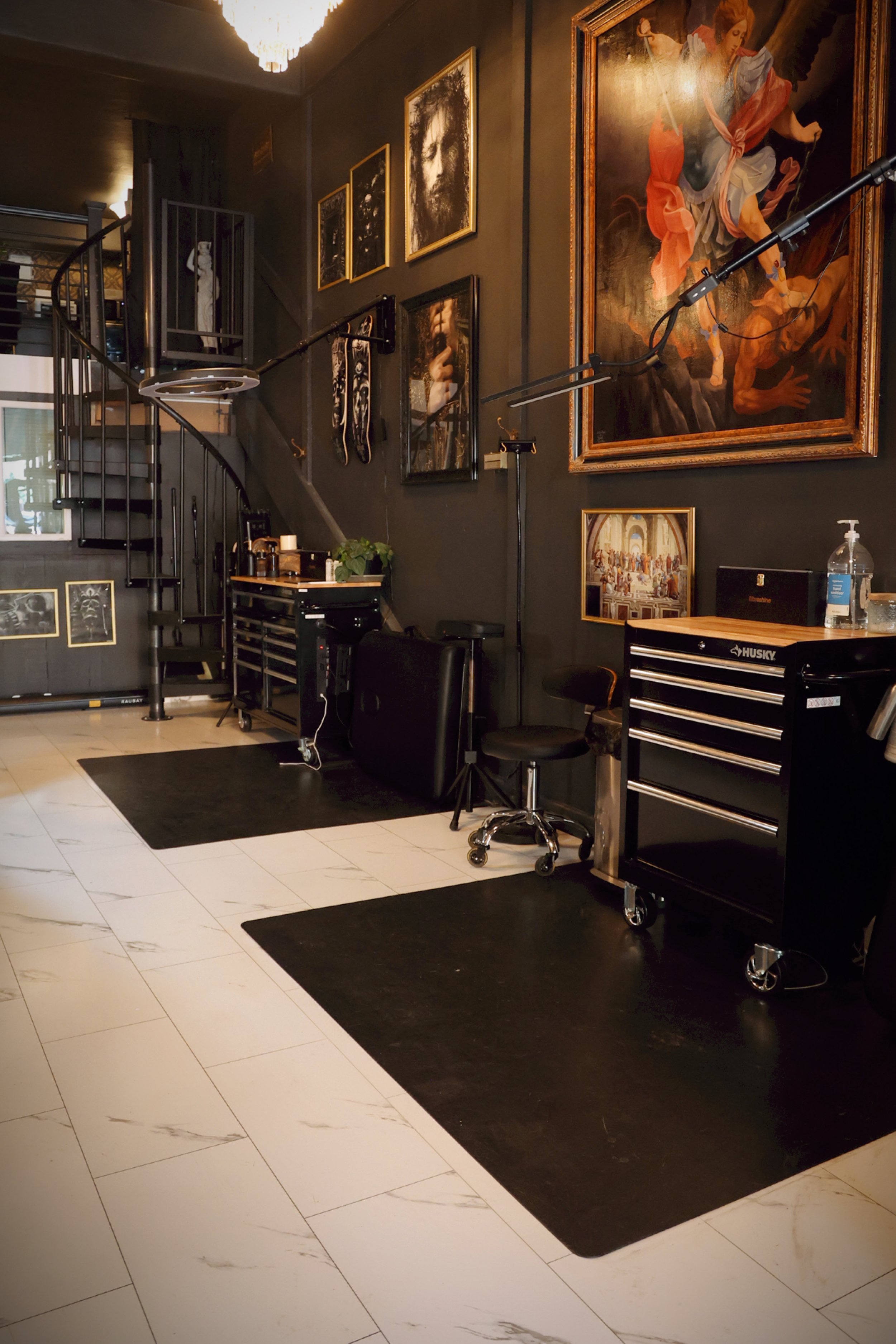 An interior of an impressive fine-art gallery in dim lighting towers over the marble floors and tattoo stations with a black spiral staircase nearing the back of the room, spiraling up to a private loft in this Boise tattoo studio. 