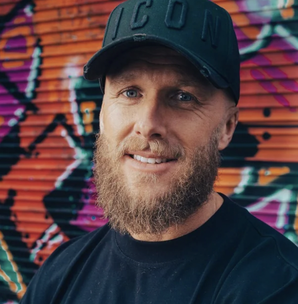 Close-up of a smiling young man with dark hair, beard, and mustache, wearing a white shirt and shoulder straps, outdoors against a light sky background.