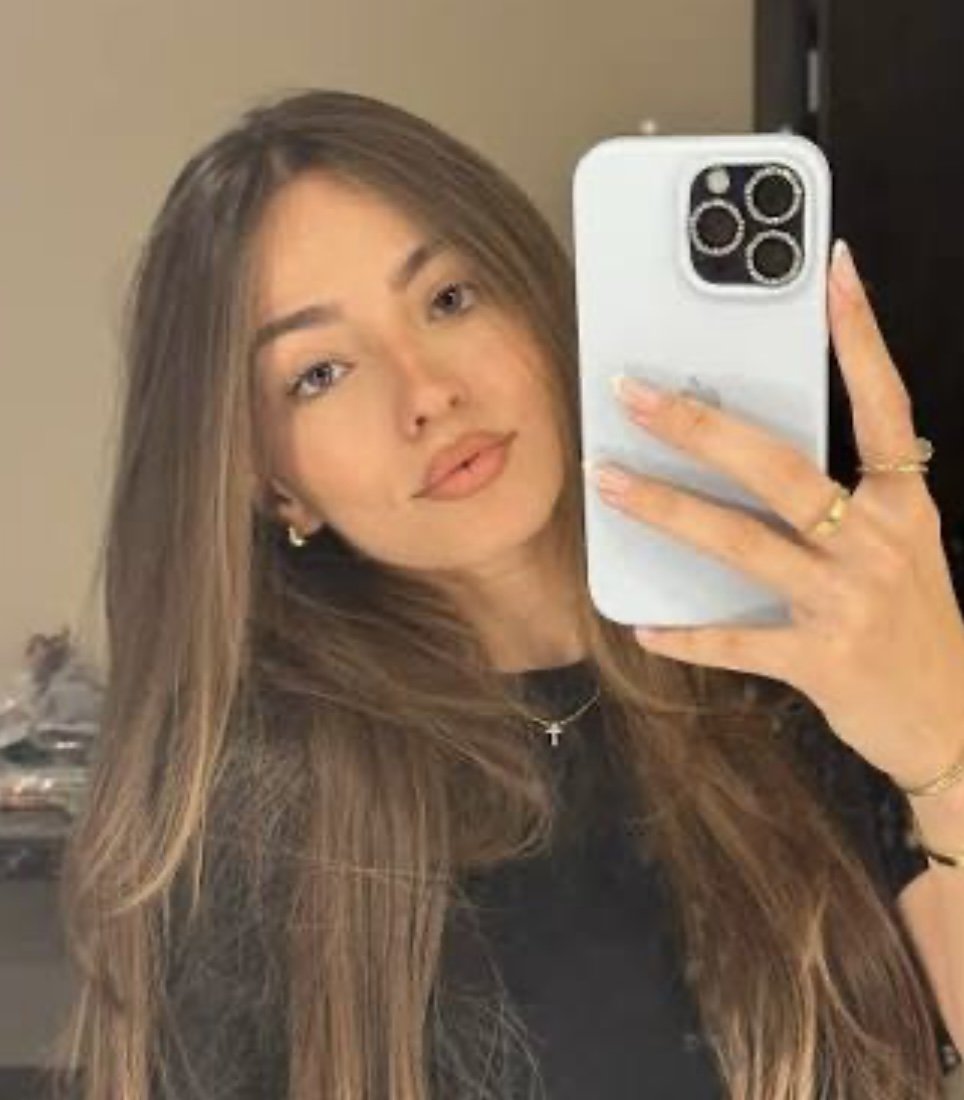 A smiling woman with short black hair, wearing a blue top, standing among lush green plants.