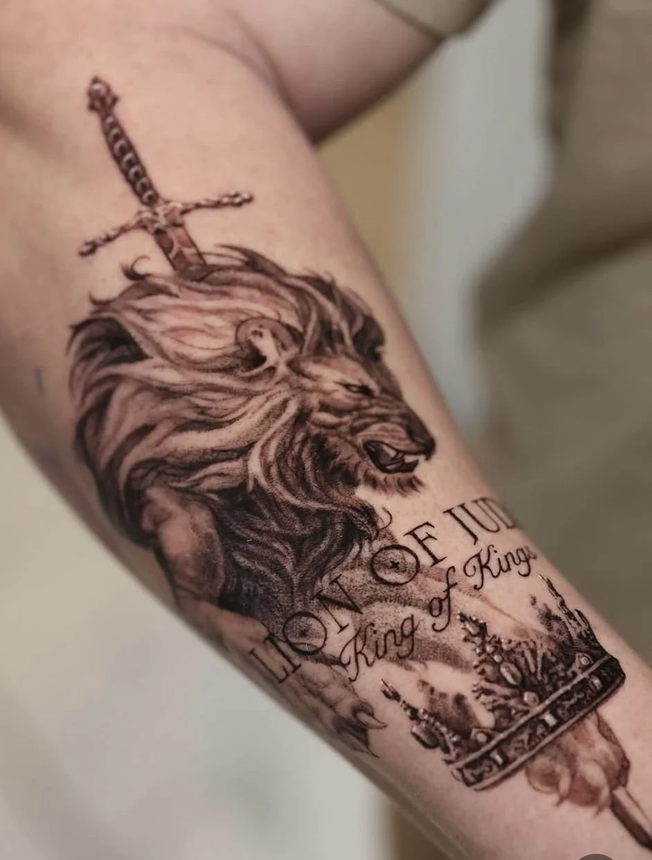 Tattoo of a lion's head with a crown and sword on a person's arm, with text reading 'Lion of Judah' and 'King of Kings' done in by the best black and gray realism tattooer in Boise, Idaho.