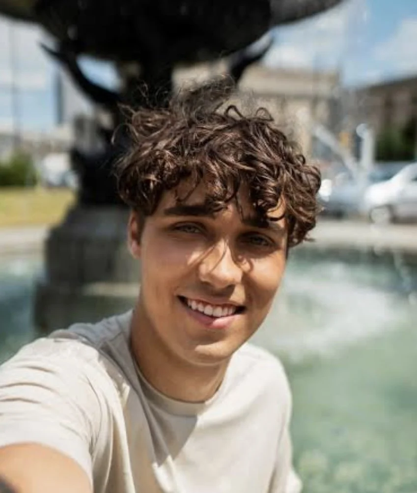 A man with curly dark hair, clear glasses, and a beard looking to the side, wearing a dark green sweater.