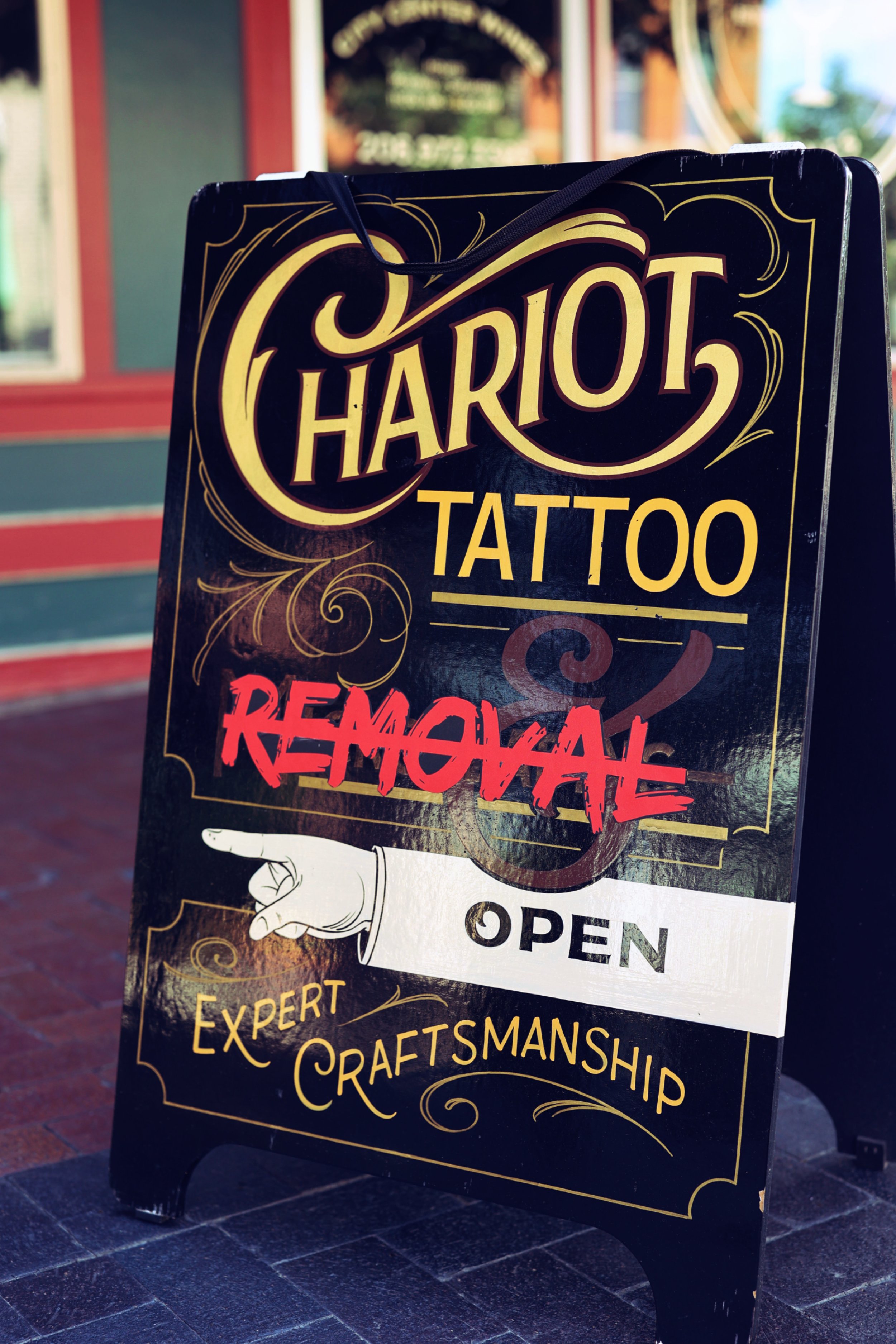 An extravagant sandwich board sign outside the studio with gold and red lettering advertising tattoos, expert craftsmanship, laser tattoo removal and open status. The sign on the sidewalk of Boise's main street points clients inside. 