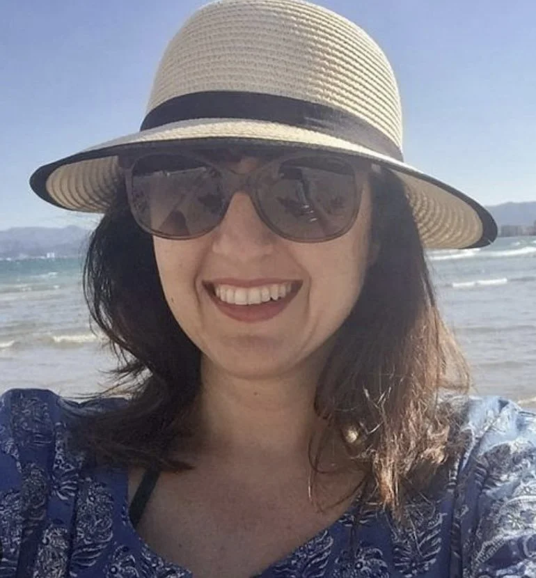 A young man wearing a black cap, sunglasses, and headphones, smiling and looking downward.
