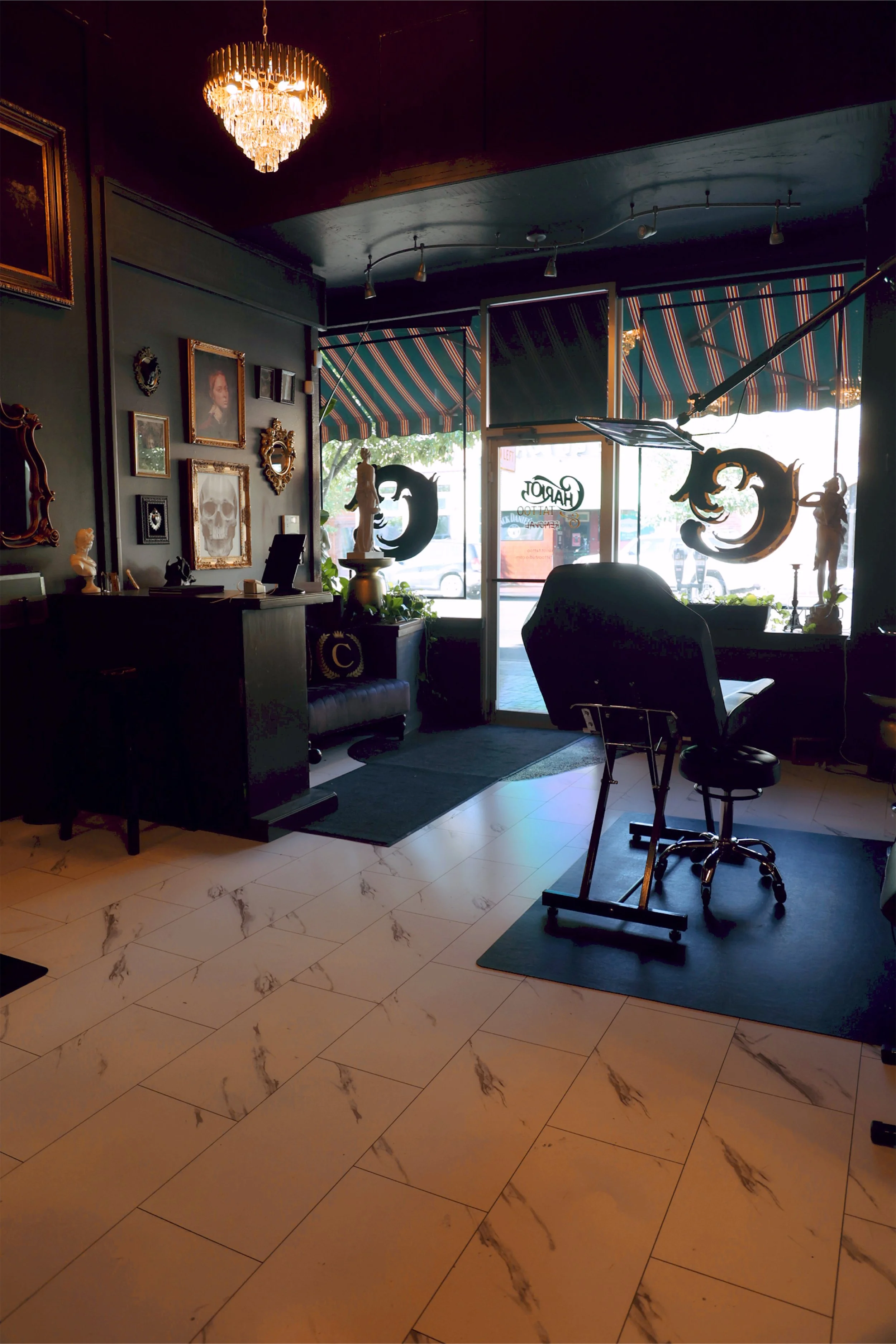 Interior of a vintage-style tattoo studio with artwork on the wall, an ornate chandelier, and tattoo chairs near the window with striped awnings outside where black and gray realism tattoo specialists shine. 