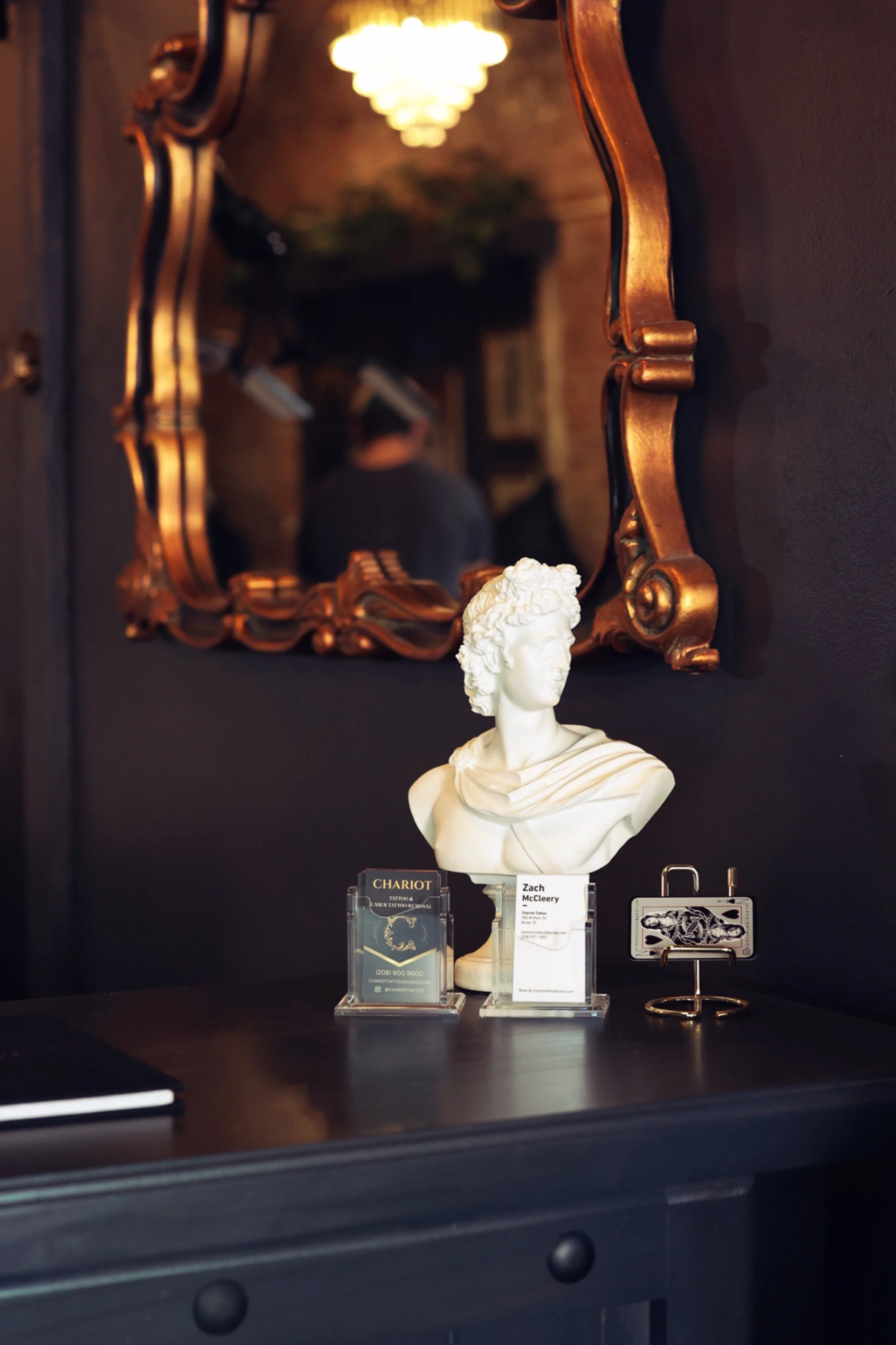 A decorative wall mirror with a golden ornate frame hangs on a dark wall, reflecting a chandelier and some plants in the background, providing a warm and decadent greeting for Boise clients entering the studio. 