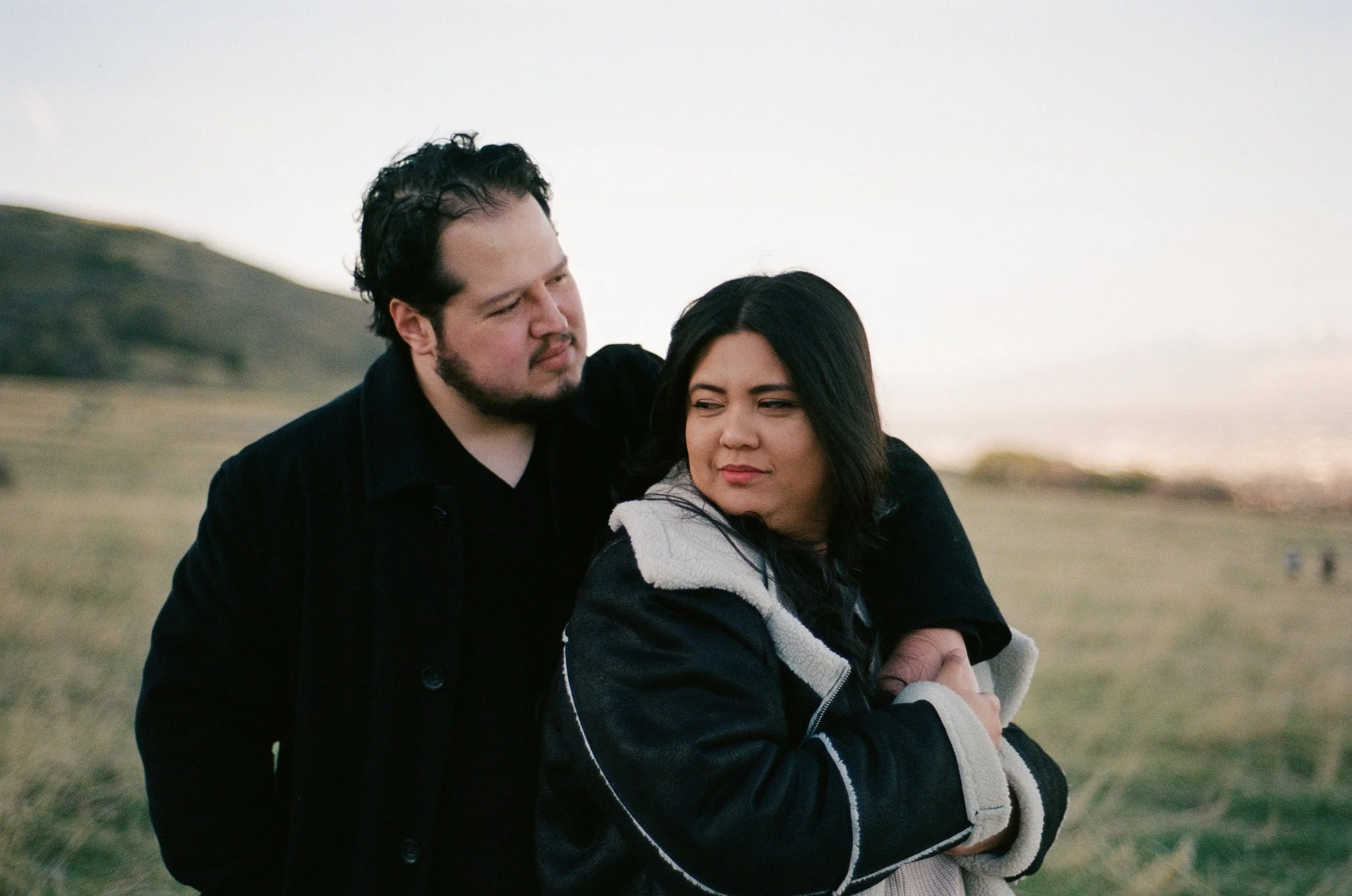 A couple stands together in a softly lit grassy field, photographed on color negative film by Poise and Ivy Imagery