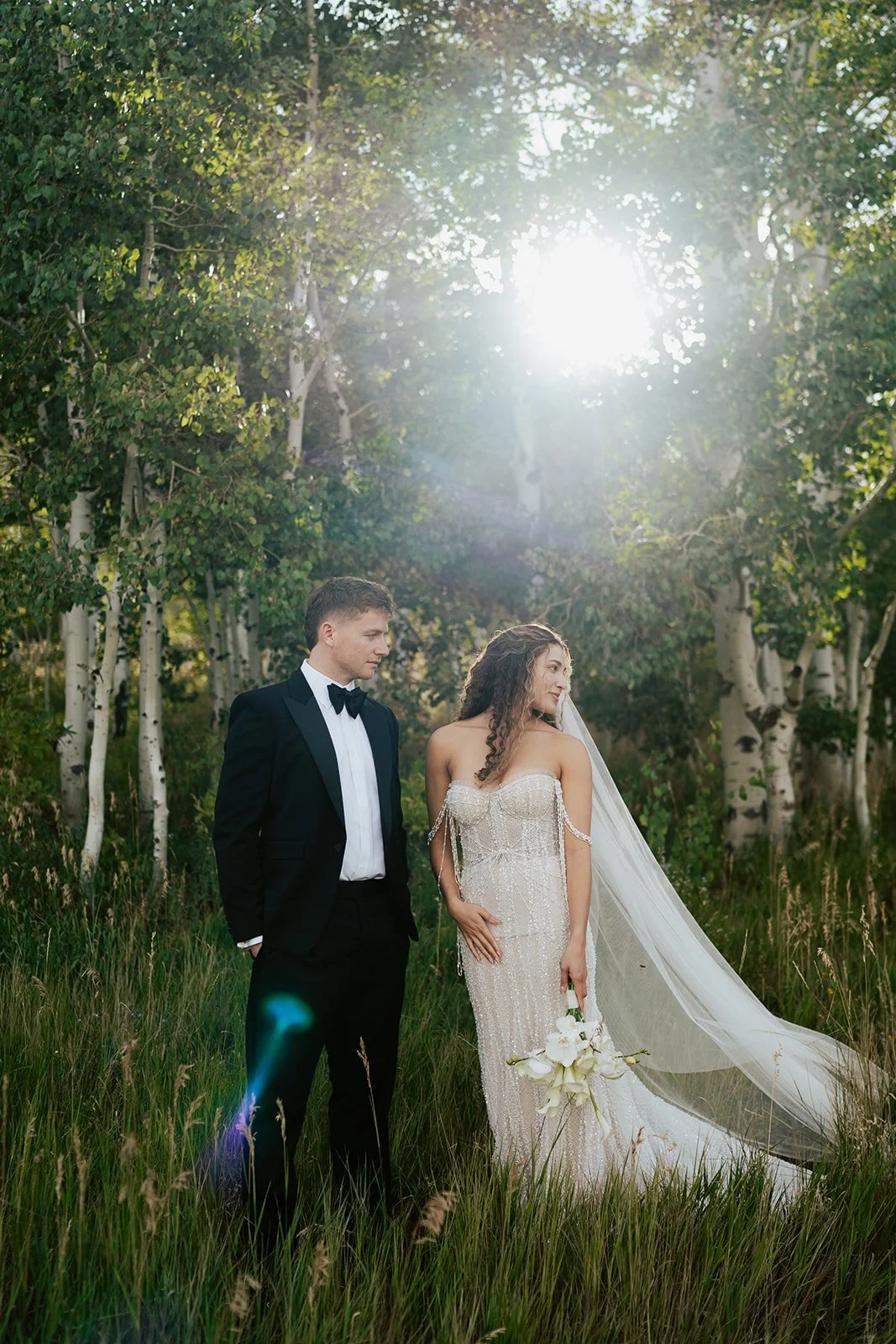 a bride and a groom stand together in a lush aspen grove. the bride wears a beaded wedding gown and the groom wears a tuxedo. Photography by Poise and Ivy Imagery