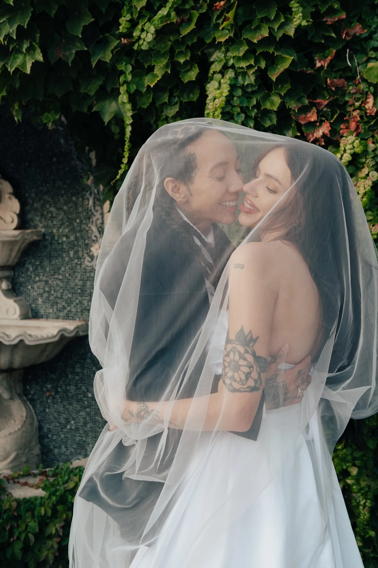 Two brides in wedding attire embrace under a wedding veil next to an ivy covered wall and an ornate fountain - photographed by Poise and Ivy Imagery
