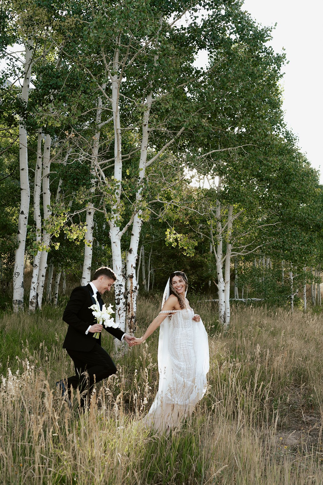 a bride and a groom stand together under a long bridal veil in a lush aspen grove. the bride wears a beaded wedding gown and the groom wears a tuxedo. Photography by Poise and Ivy Imagery