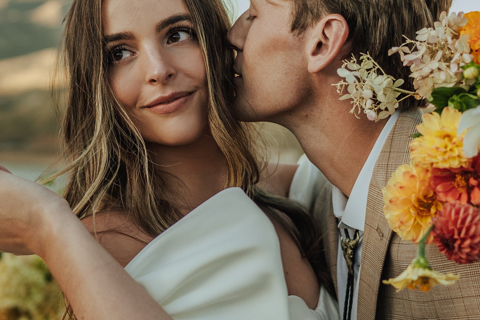 A bride and groom in a wedding gown and a tan suit stand in a meadow with mountains and a lake behind them - photography by Poise and Ivy Imagery