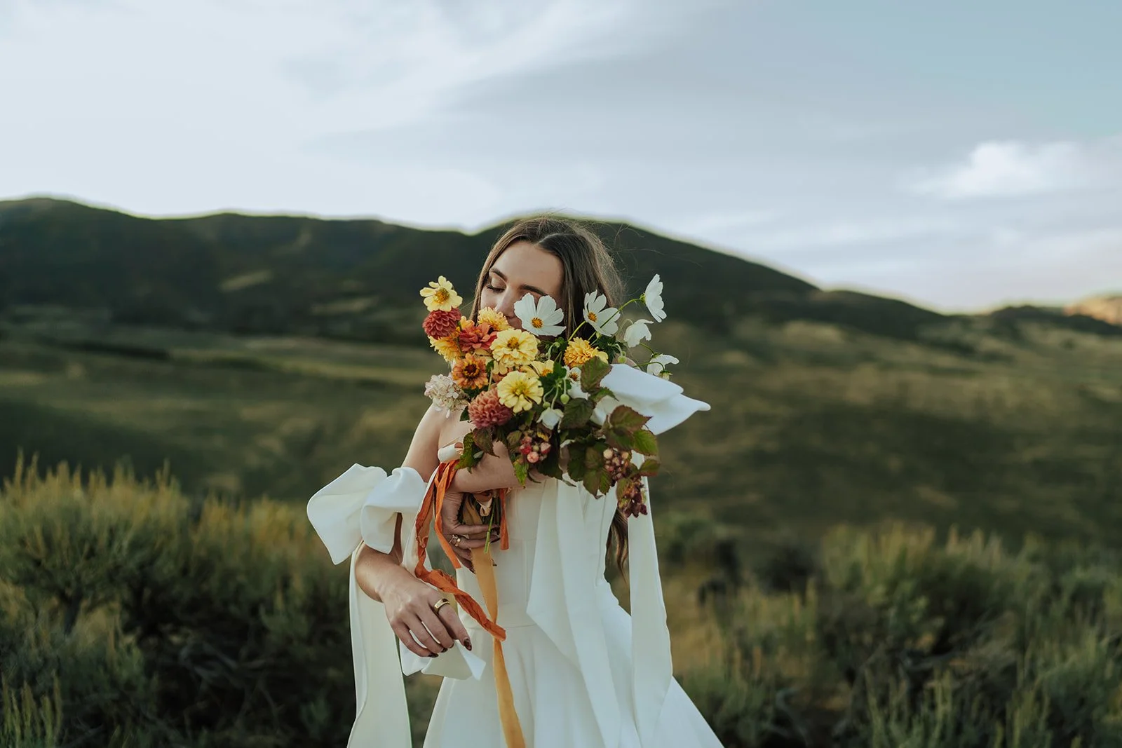 A bride in a wedding gown stands in a meadow with mountains behind her, holding her warm toned wedding bouquet - photography by Poise and Ivy Imagery