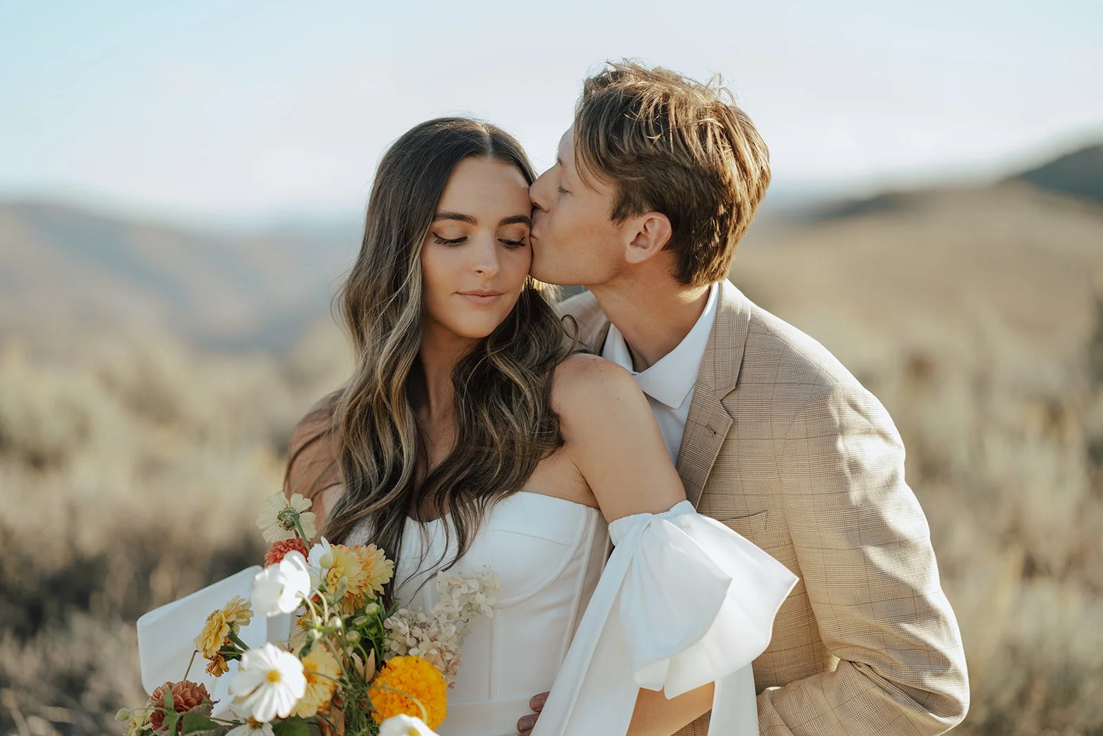 A bride and groom in a wedding gown and a tan suit stand in a meadow with mountainous peaks behind them - photography by Poise and Ivy Imagery
