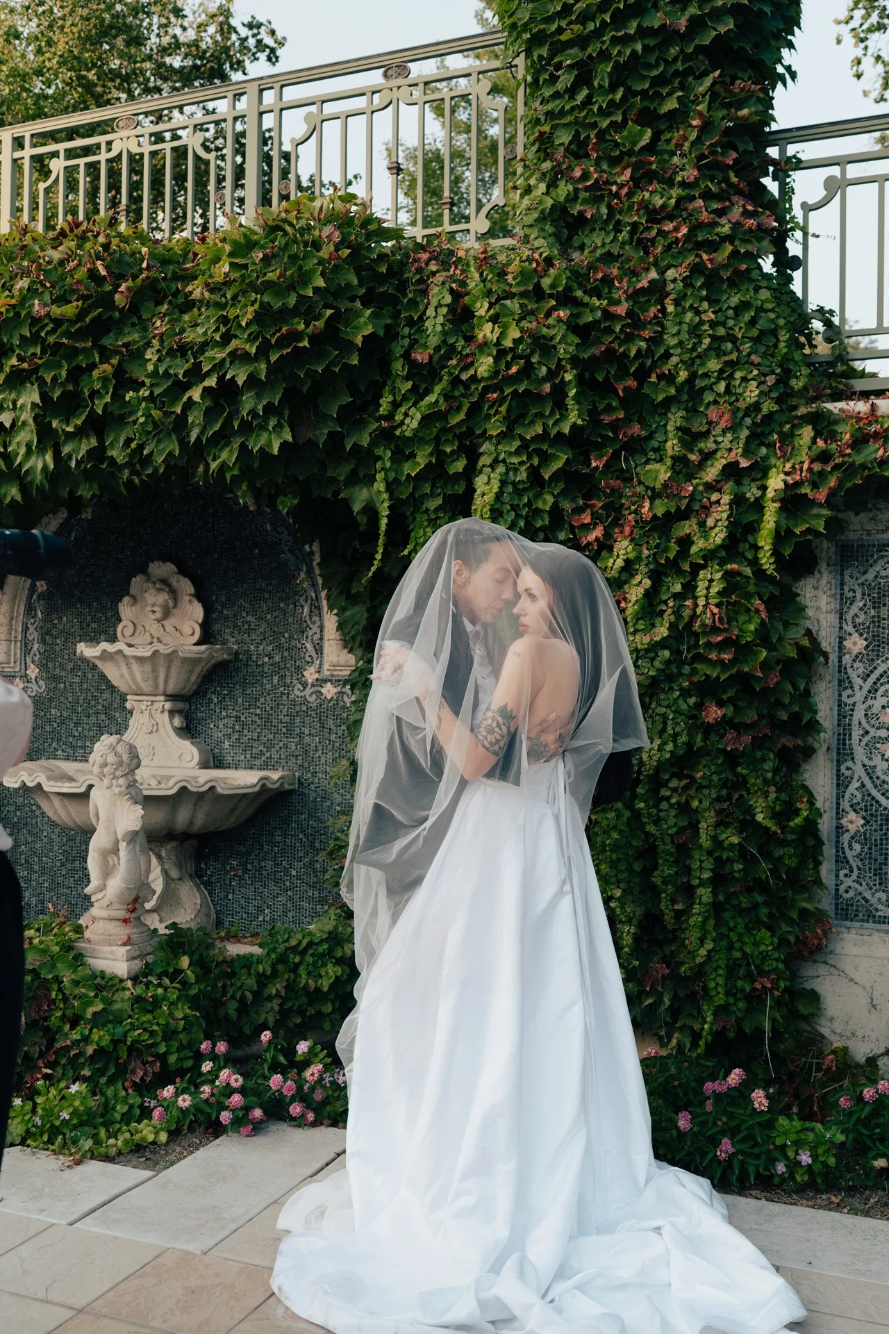 Two brides in wedding attire embrace under an ornate outdoor arched corridor - photographed by Poise and Ivy Imagery