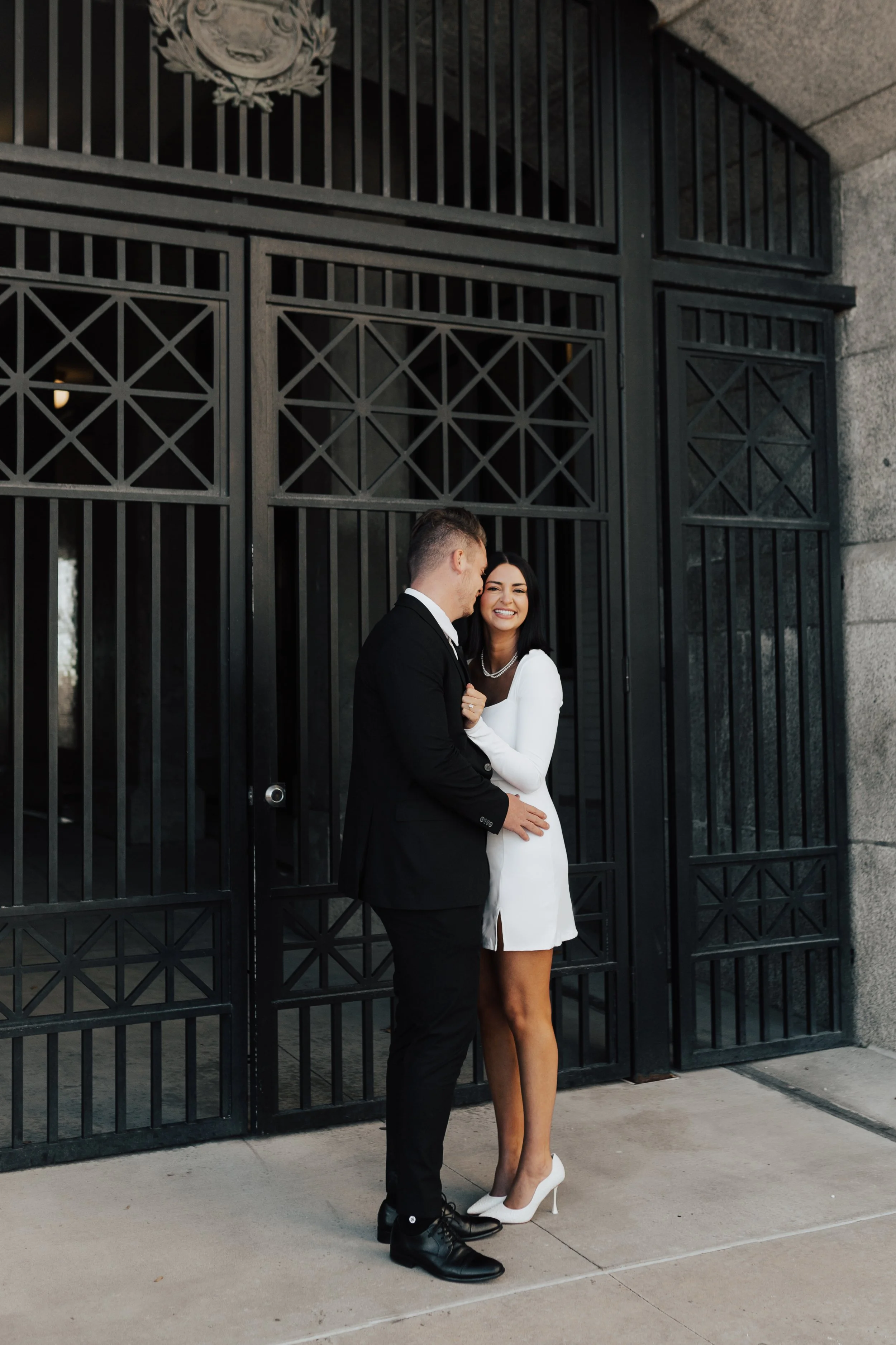 A couple standing together against a black wrought iron gate wearing a white dress and a black suit. Couple's photoshoot - Poise and Ivy Imagery