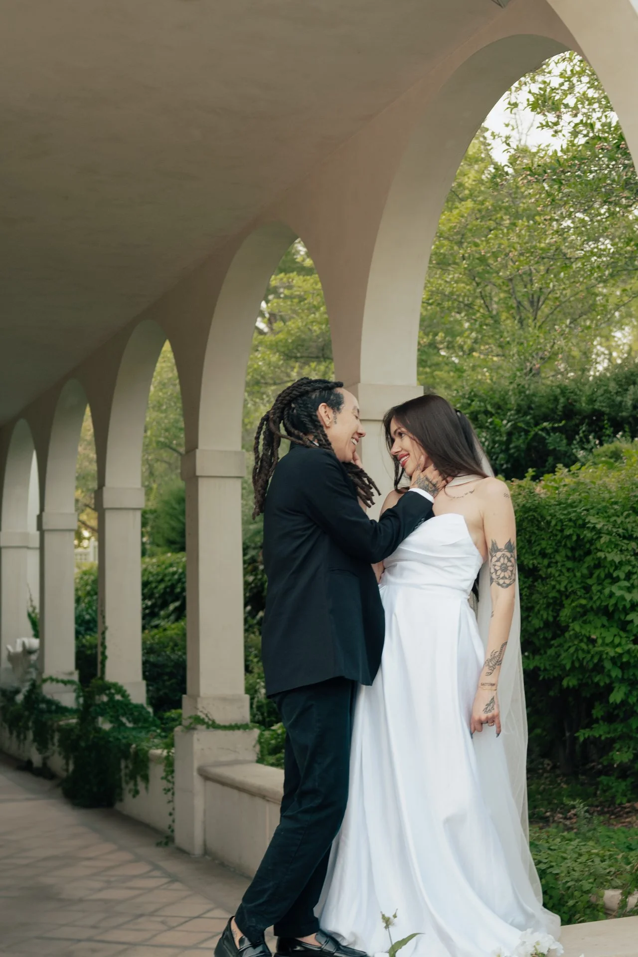 Two brides in wedding attire embrace under an ornate outdoor arched corridor - photographed by Poise and Ivy Imagery