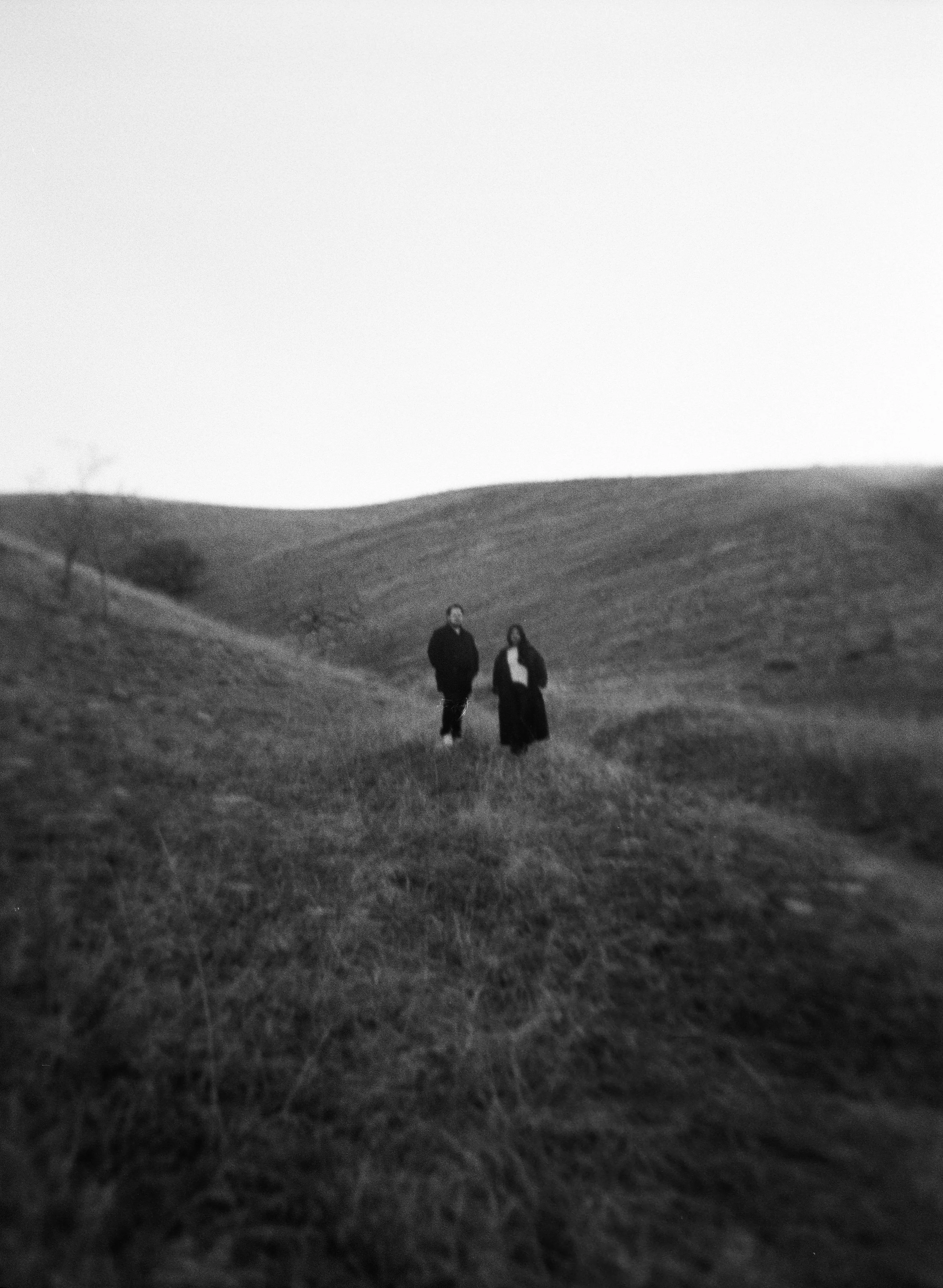 A couple stands together in a softly lit grassy field on black and white medium format film - photography by Poise and Ivy Imagery