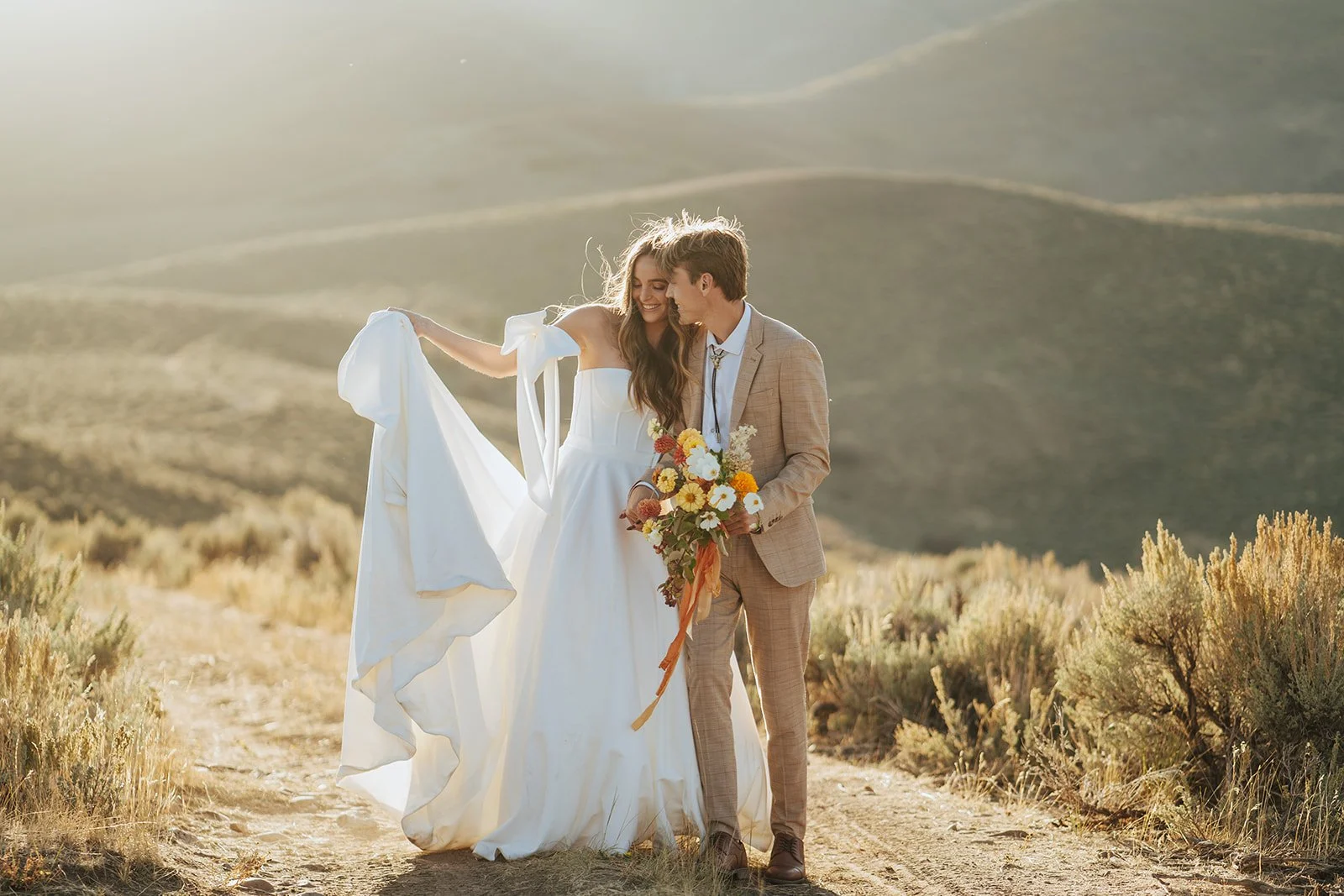 A bride and groom in a wedding gown and a tan suit stand in a meadow with mountainous peaks behind them - photography by Poise and Ivy Imagery