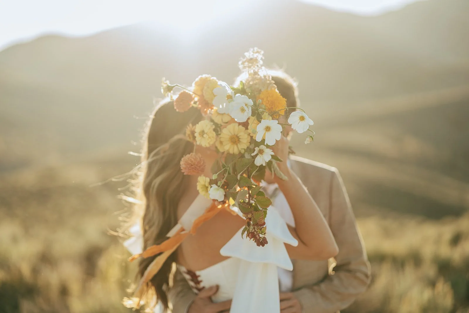A bride and groom in a wedding gown and a tan suit stand in a meadow with mountainous peaks behind them - photography by Poise and Ivy Imagery