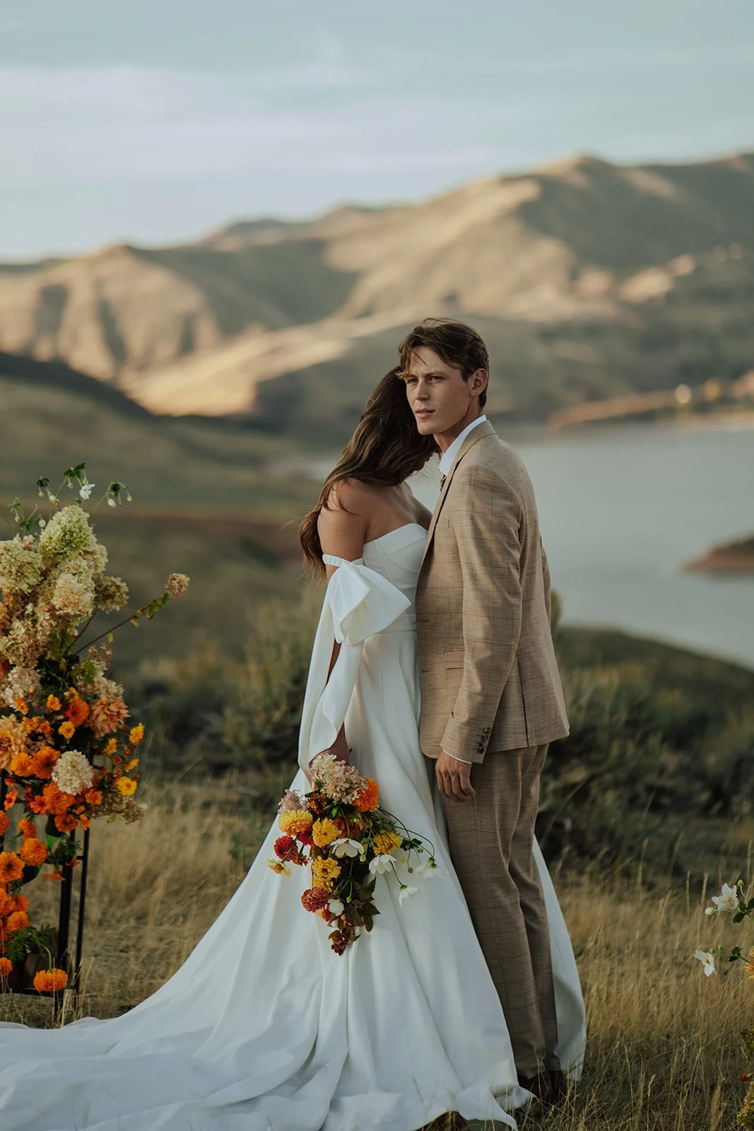 A bride and groom in a wedding gown and a tan suit stand in a meadow with mountains and a lake behind them - photography by Poise and Ivy Imagery