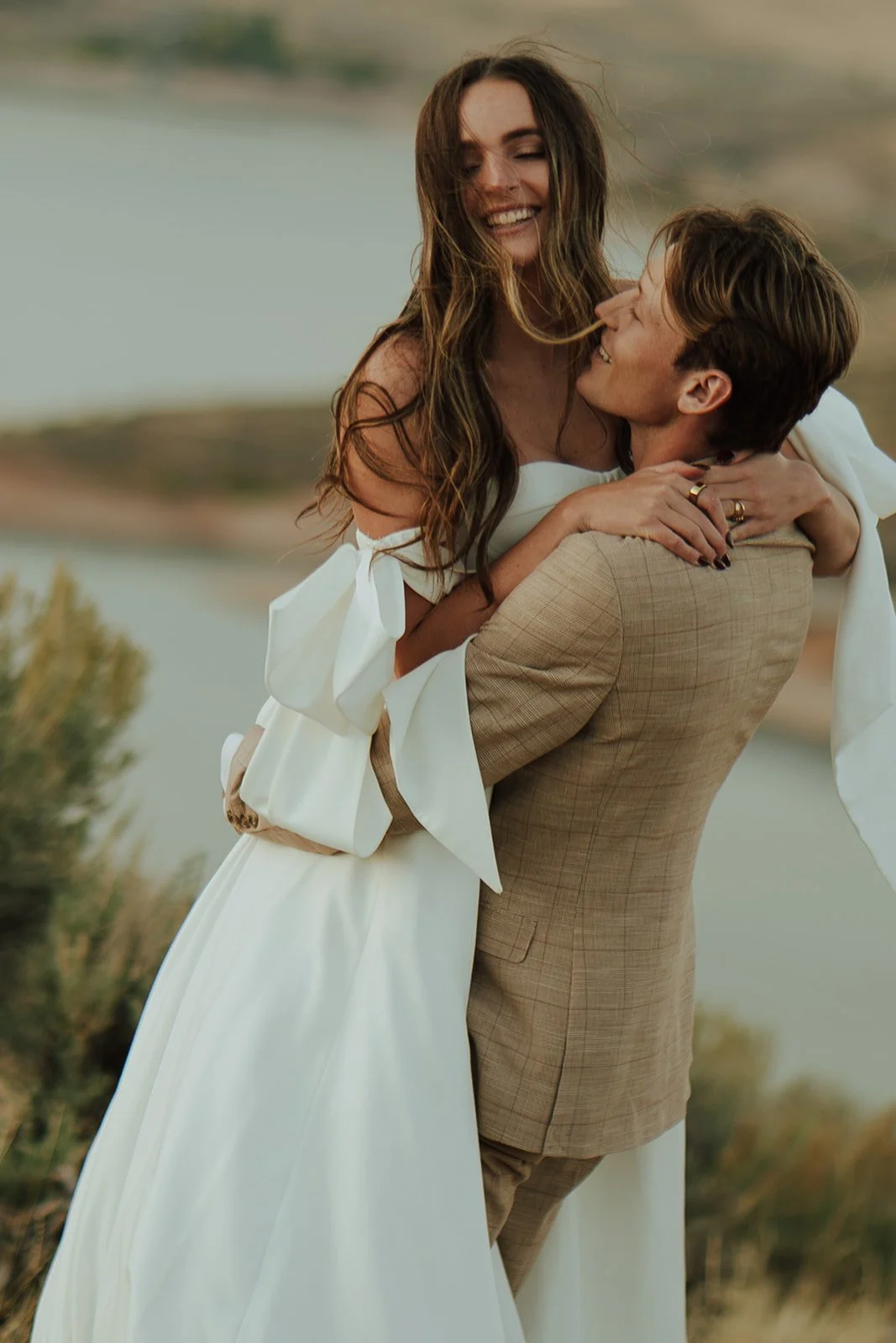 A bride and groom in a wedding gown and a tan suit stand in a meadow with mountains and a lake behind them - photography by Poise and Ivy Imagery