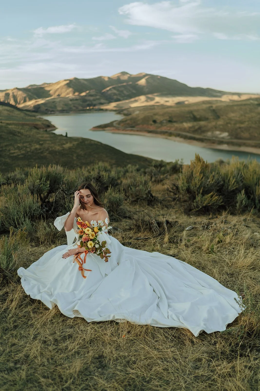 A bride sits in a meadow in her wedding dress overlooking mountains. photography by Poise and Ivy Imagery