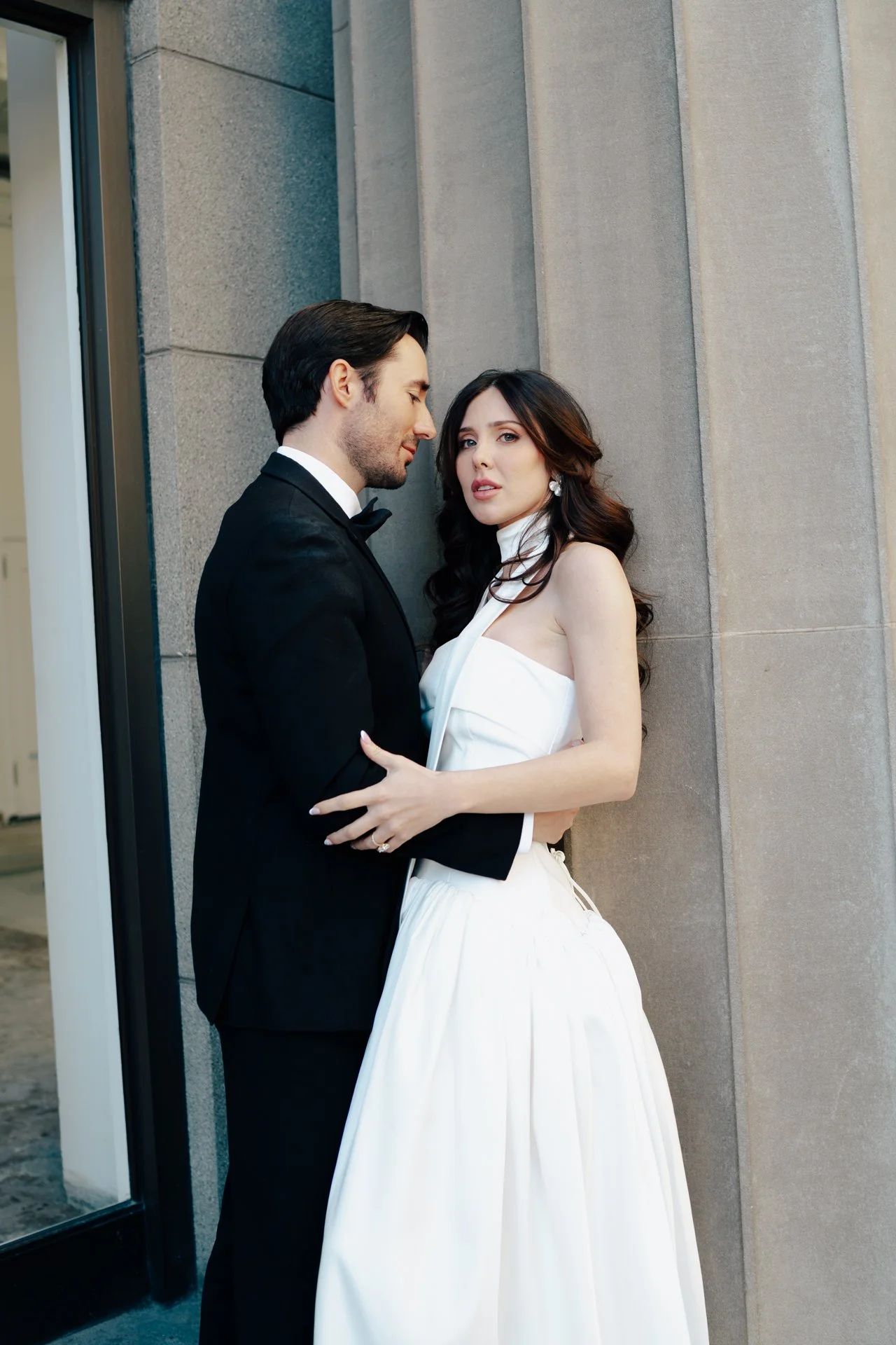 A bride and groom stand together among roman - style architecture in an elegant wedding gown and a classic black suit