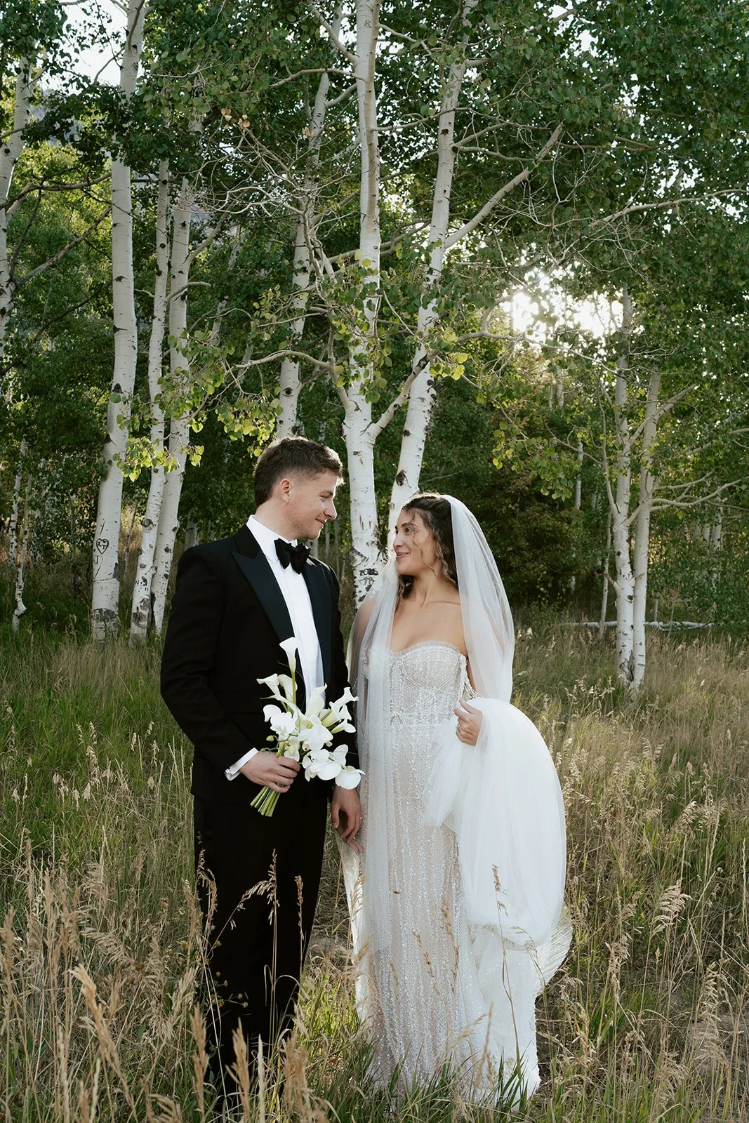 a bride and a groom stand together under a long bridal veil in a lush aspen grove. the bride wears a beaded wedding gown and the groom wears a tuxedo. Photography by Poise and Ivy Imagery