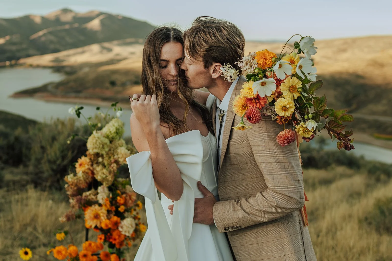 A bride and groom in a wedding gown and a tan suit stand in a meadow with mountains and a lake behind them - photography by Poise and Ivy Imagery