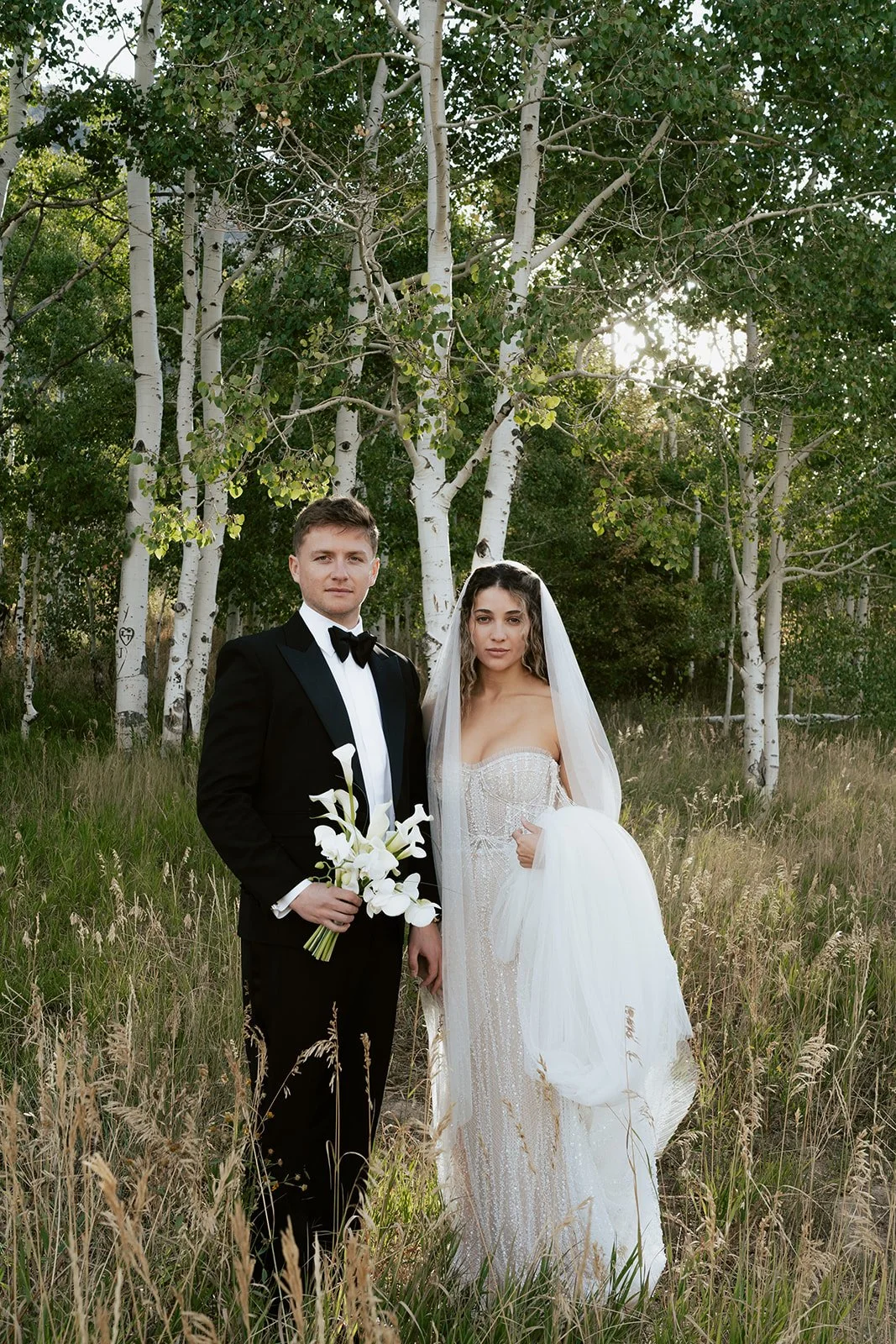 a bride and a groom walk together in a lush aspen grove. the bride wears a beaded wedding gown with a bridal veil and the groom wears a tuxedo. Photography by Poise and Ivy Imagery