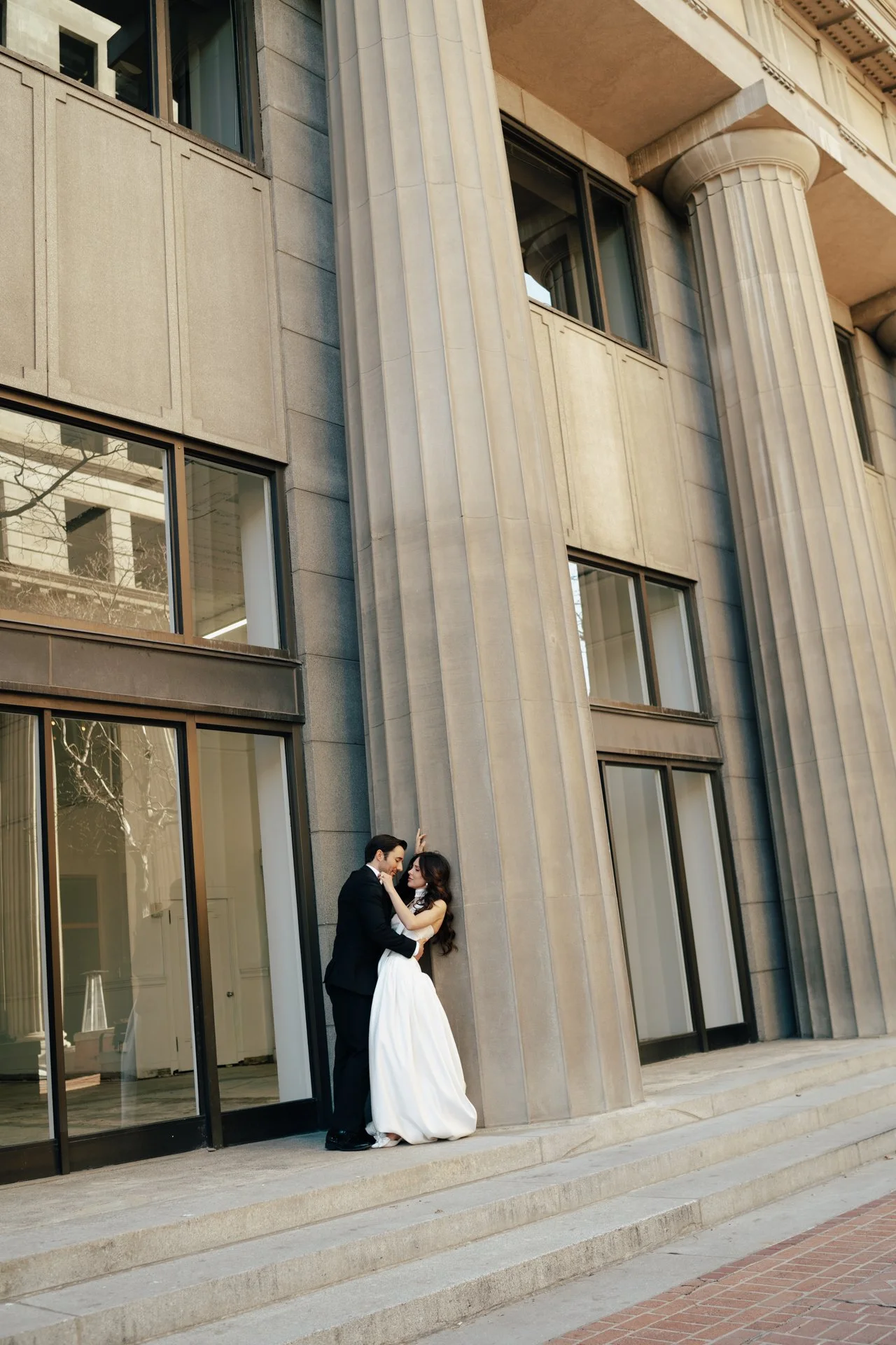 A bride and groom stand together among roman - style architecture in an elegant wedding gown and a classic black suit