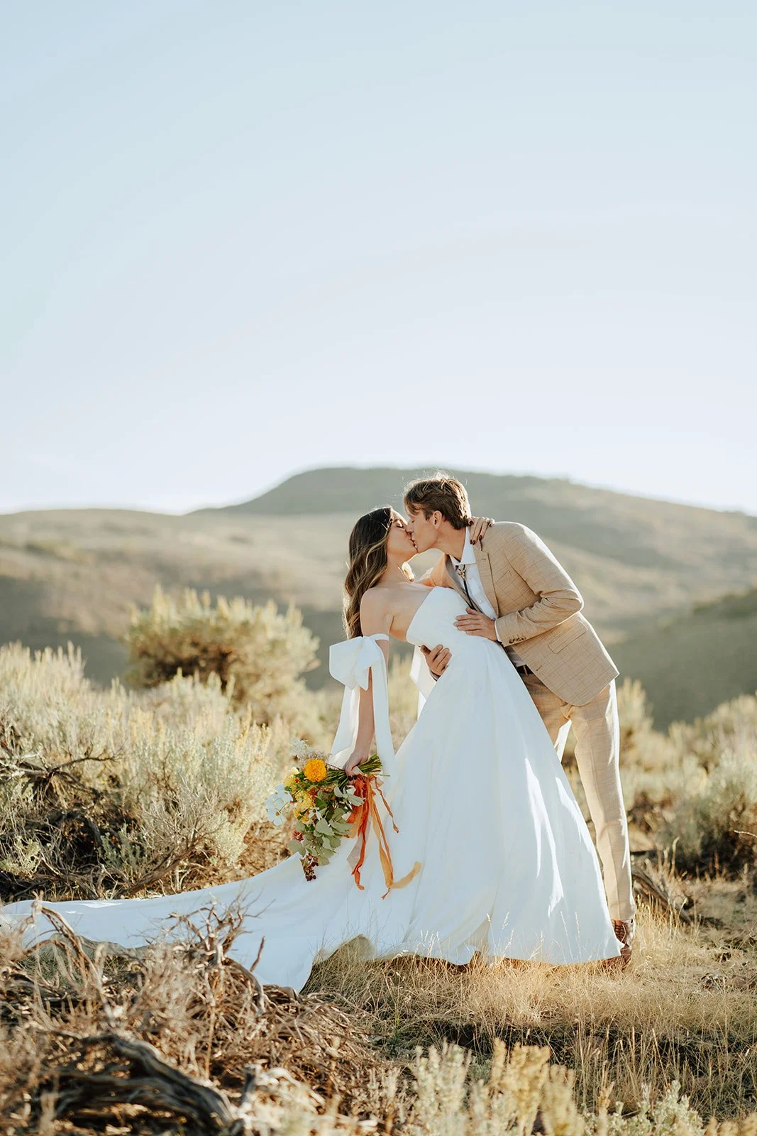 A bride and groom in a wedding gown and a tan suit stand in a meadow with mountainous peaks behind them - photography by Poise and Ivy Imagery