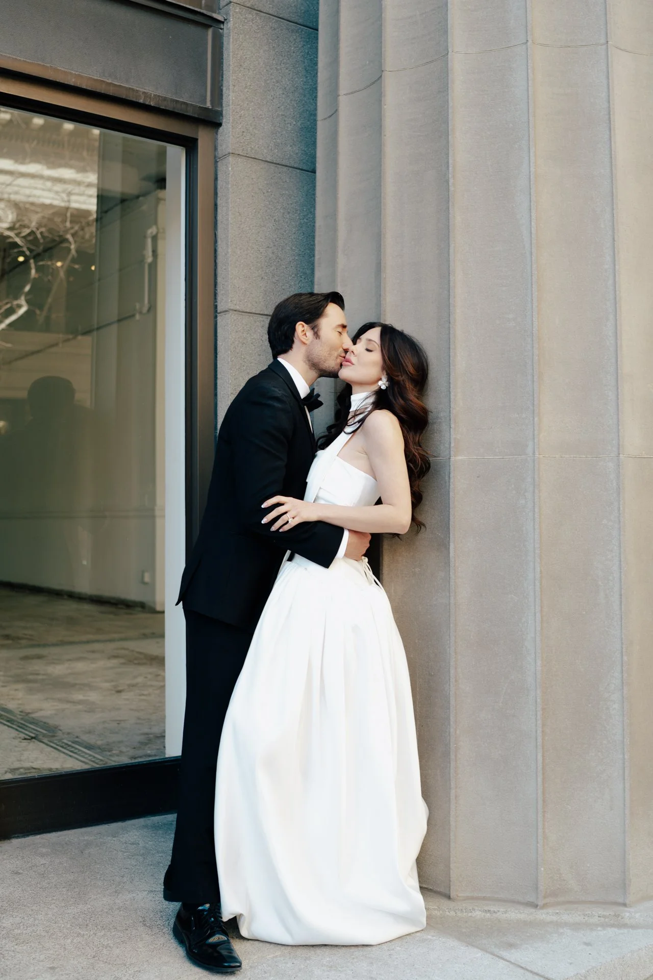 A bride and groom stand together among roman - style architecture in an elegant wedding gown and a classic black suit