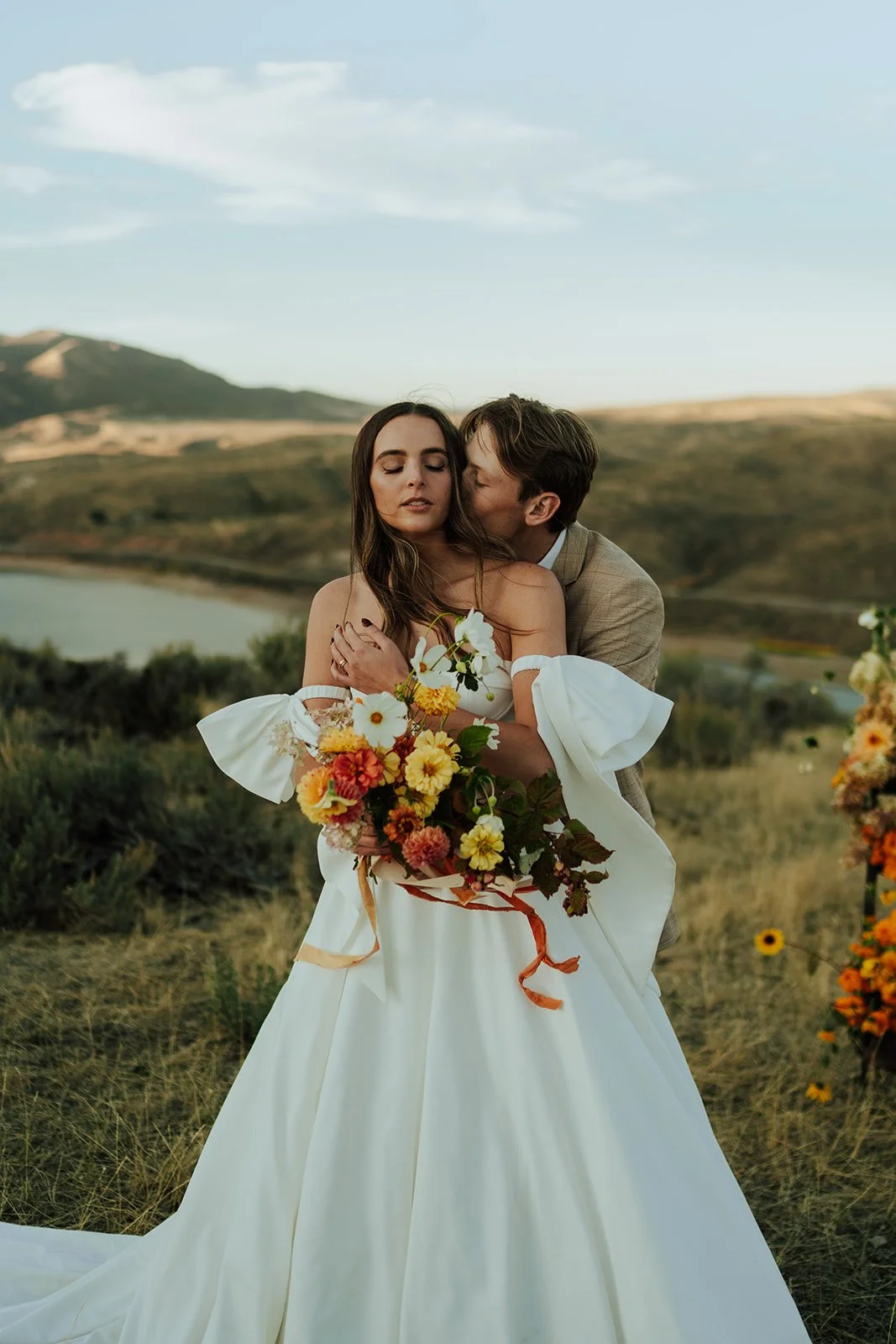 A bride and groom in a wedding gown and a tan suit stand in a meadow with mountains and a lake behind them - photography by Poise and Ivy Imagery