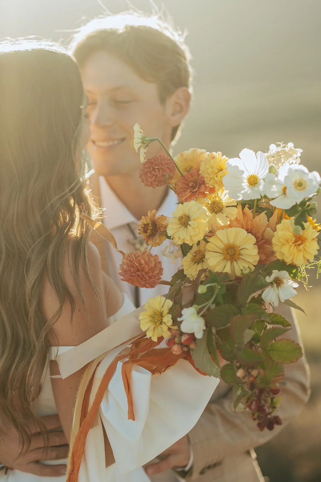 A bride and groom in a wedding gown and a tan suit stand in a meadow with mountainous peaks behind them - photography by Poise and Ivy Imagery