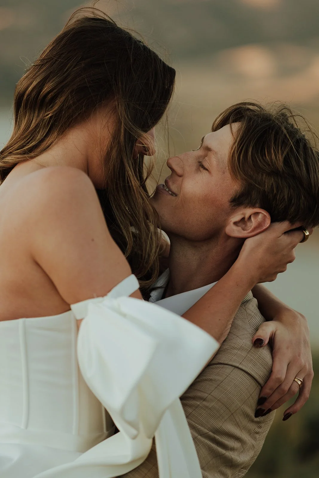 A bride and groom in a wedding gown and a tan suit stand in a meadow with mountains and a lake behind them - photography by Poise and Ivy Imagery