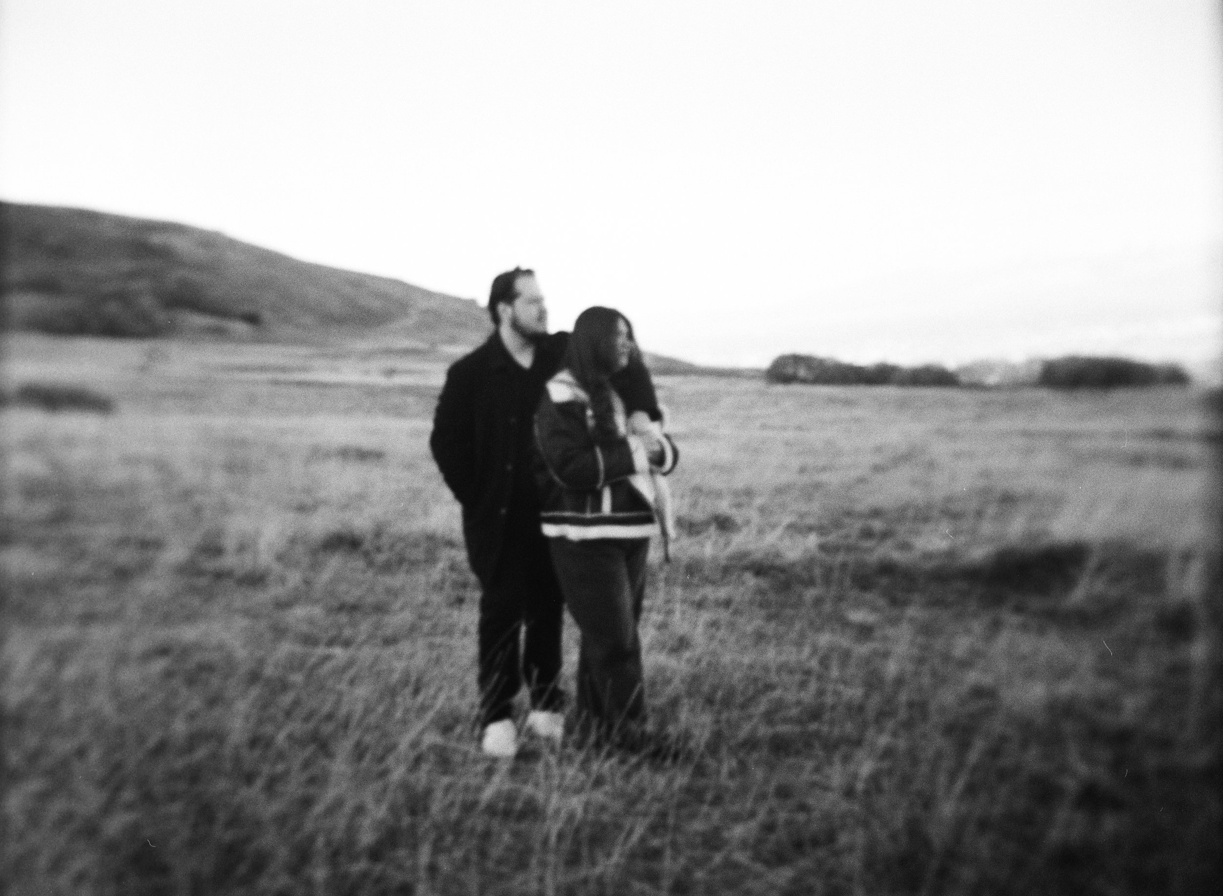 A couple stands together in a softly lit grassy field on black and white medium format film - photography by Poise and Ivy Imagery