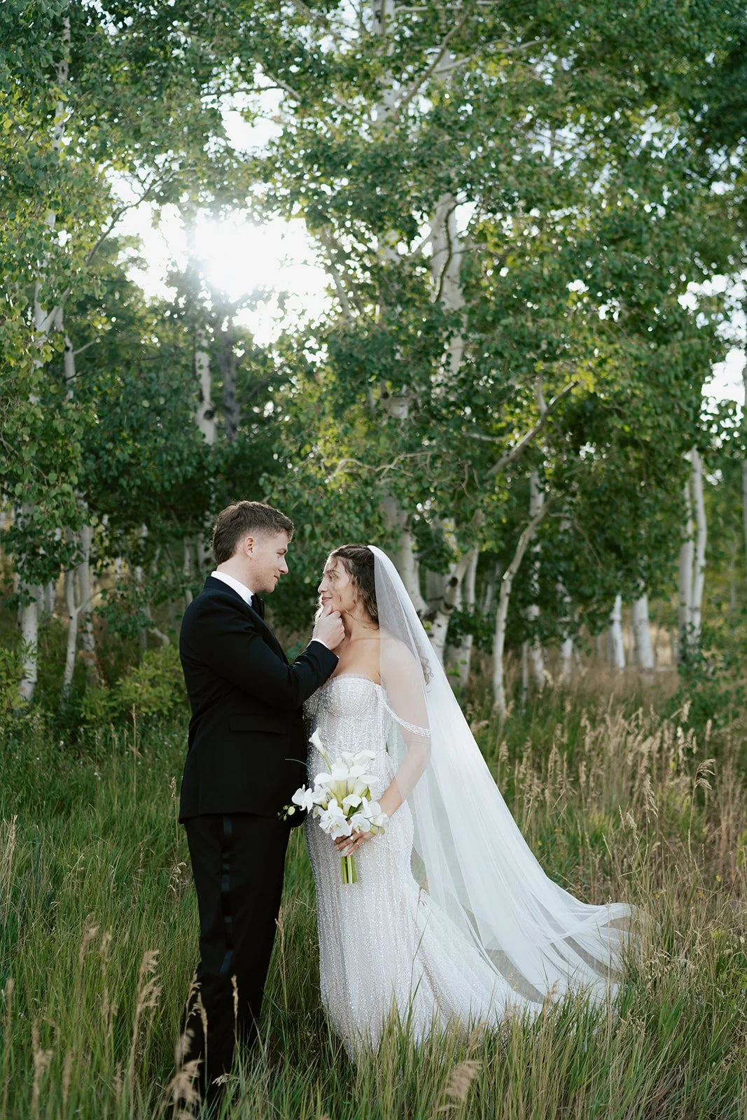 a bride and a groom stand together in a lush aspen grove. the bride wears a beaded wedding gown and the groom wears a tuxedo. Photography by Poise and Ivy Imagery