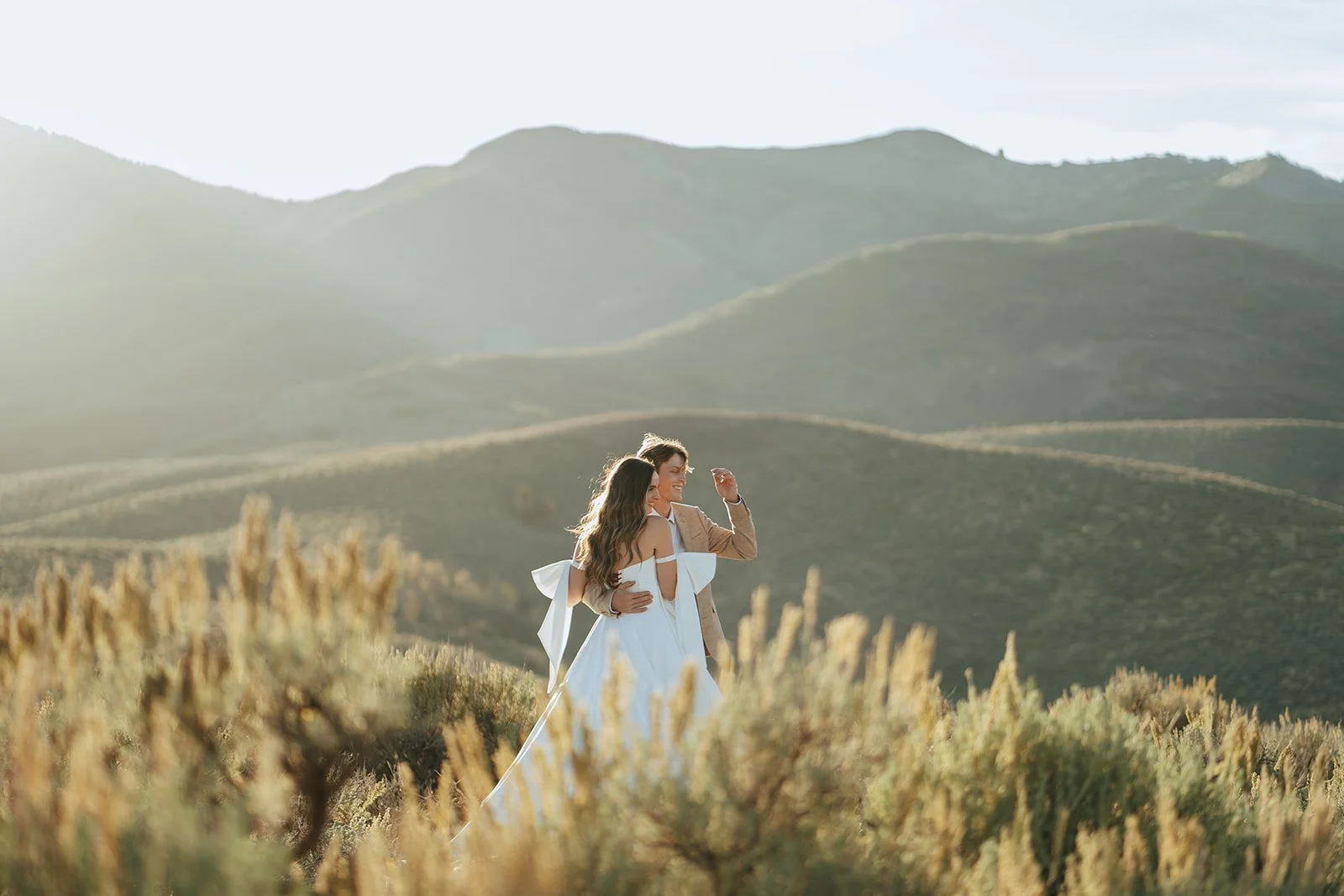 A bride and groom in a wedding gown and a tan suit stand in a meadow with mountainous peaks behind them - photography by Poise and Ivy Imagery