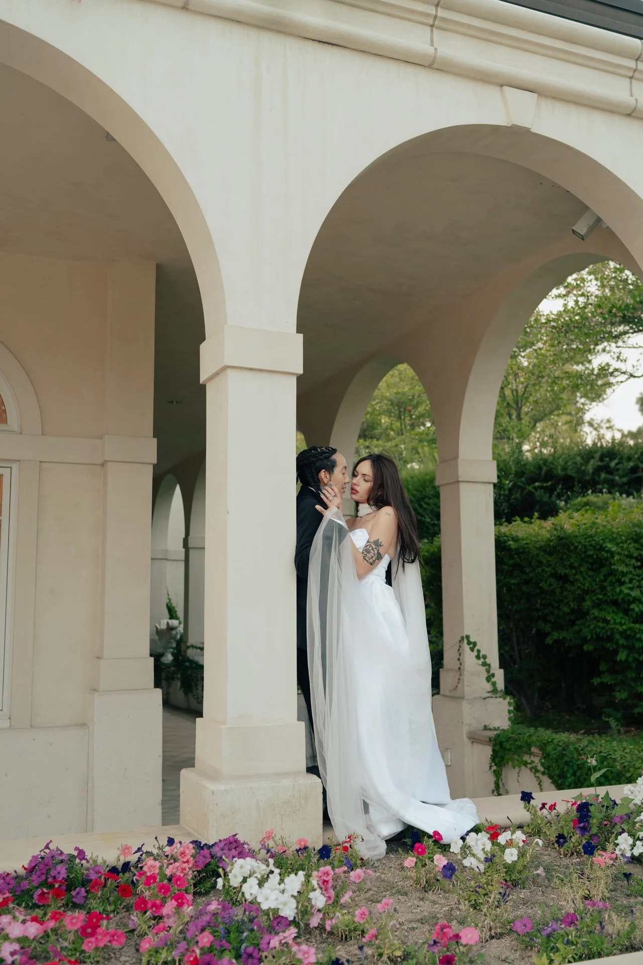 Two brides in wedding attire embrace under an ornate outdoor arched corridor - photographed by Poise and Ivy Imagery
